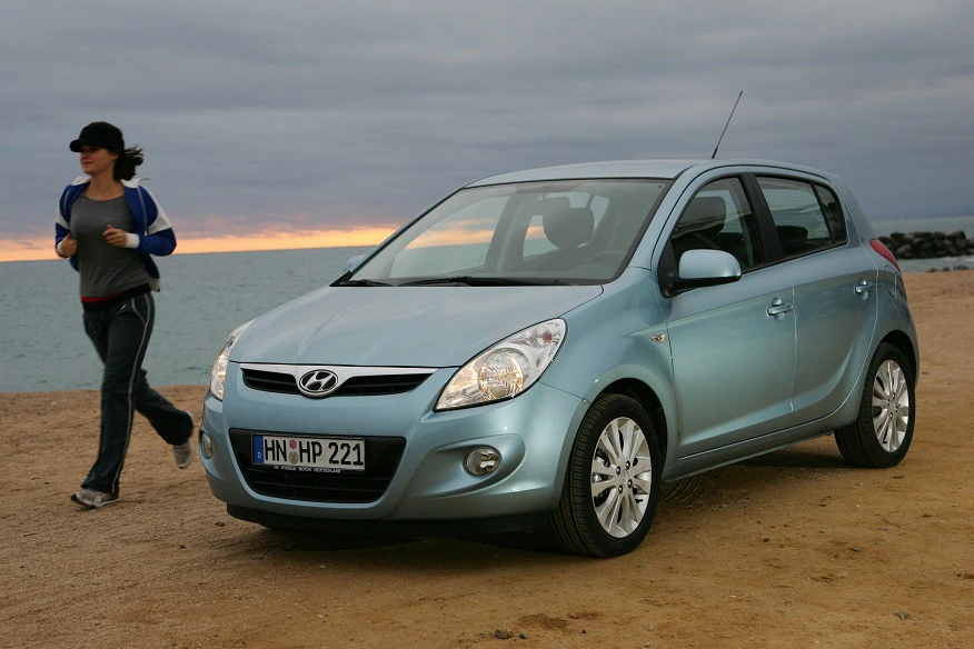 Ein blau-silberner Hyundai i20 steht am Strand. Im Hintergrund joggt eine Person entlang der Küste bei bewölktem Himmel.