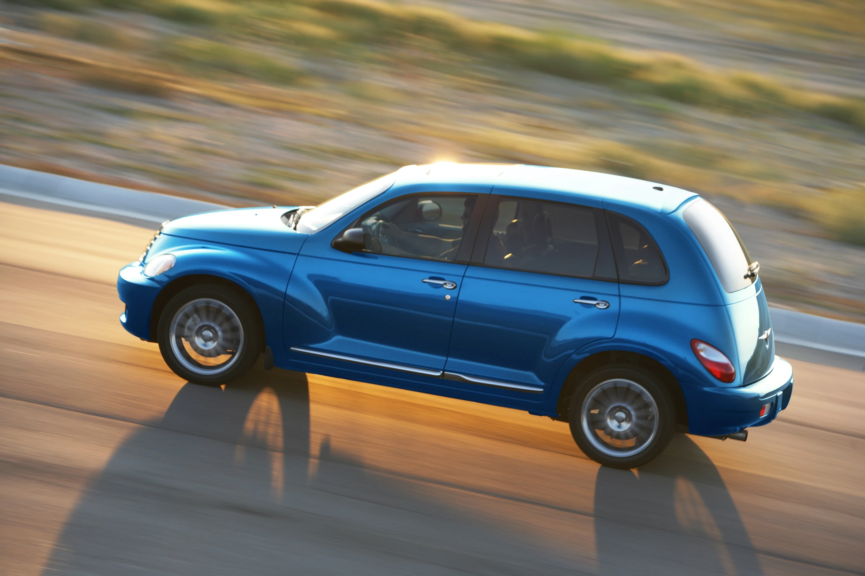 Blauer Chrysler PT Cruiser fährt auf einer Straße in Bewegung, mit Sonnenlicht, das auf die Karosserie scheint.