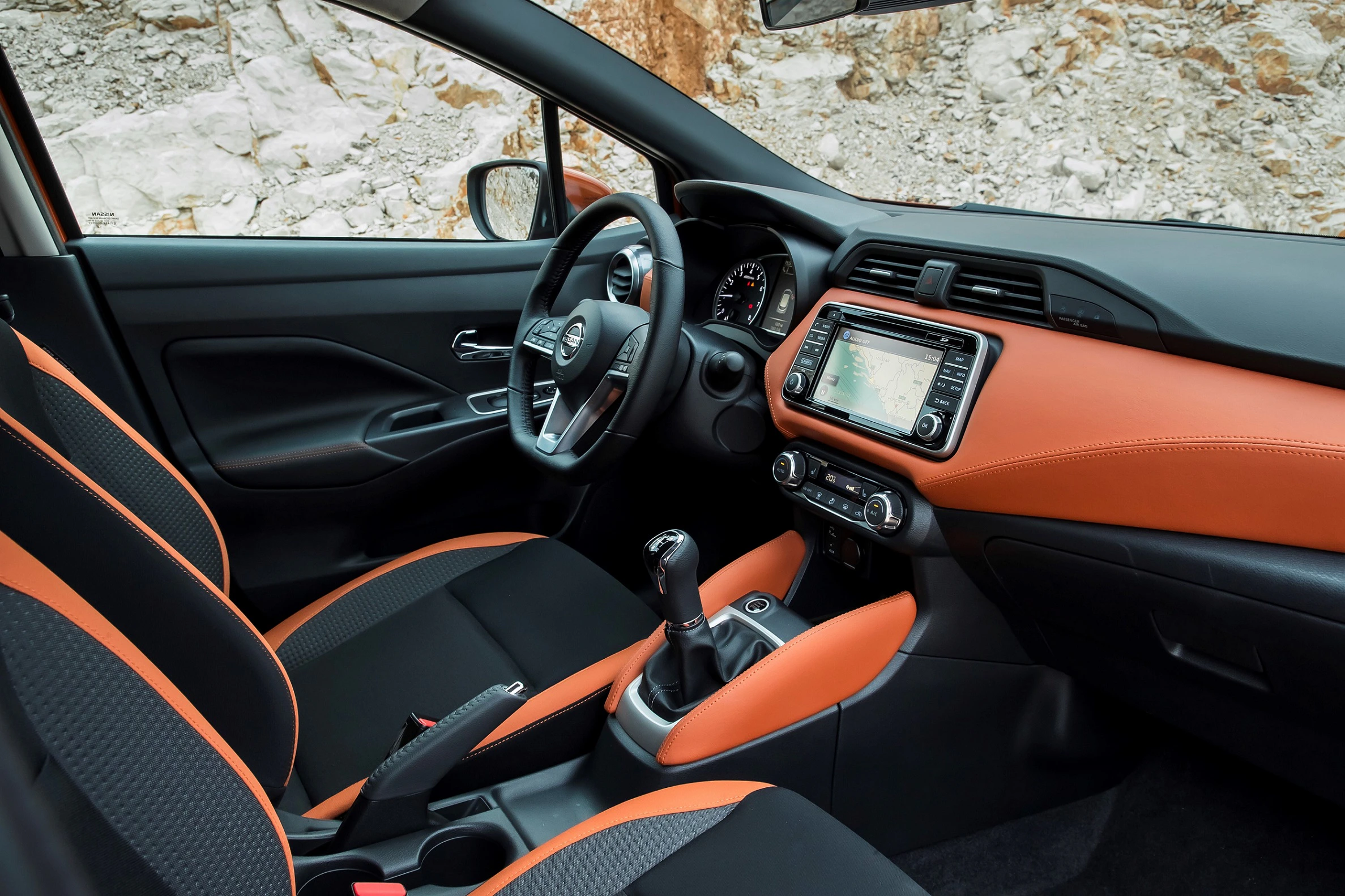 Modernes Auto-Interieur mit orange-schwarzen Sitzen, Lenkrad, und eingebautem Display vor einer felsigen Kulisse.