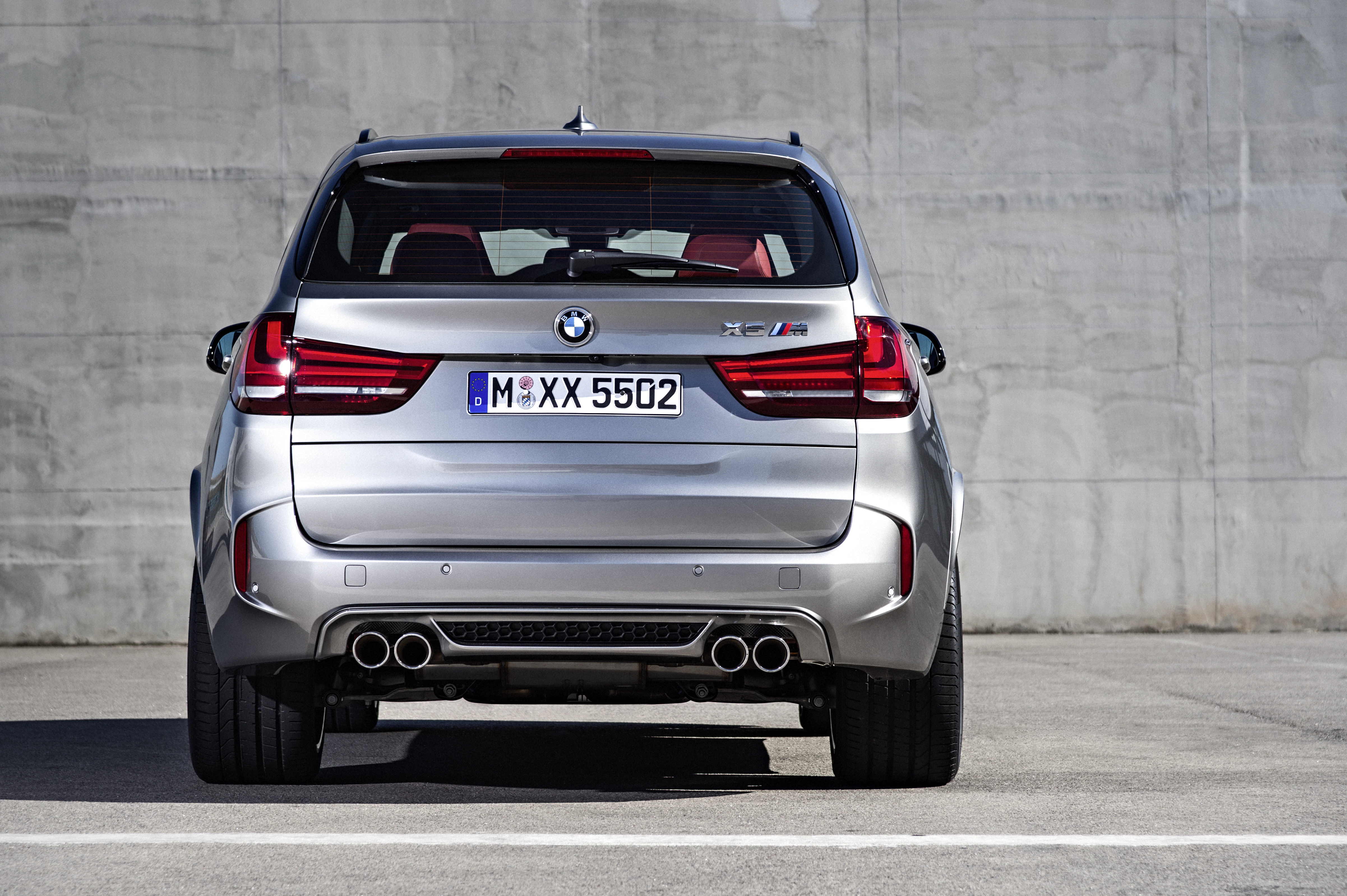Silberner BMW X5 M, Heckansicht vor grauer Betonwand, mit sichtbaren Auspuffrohren und Leuchten, roter Innenraum erkennbar.