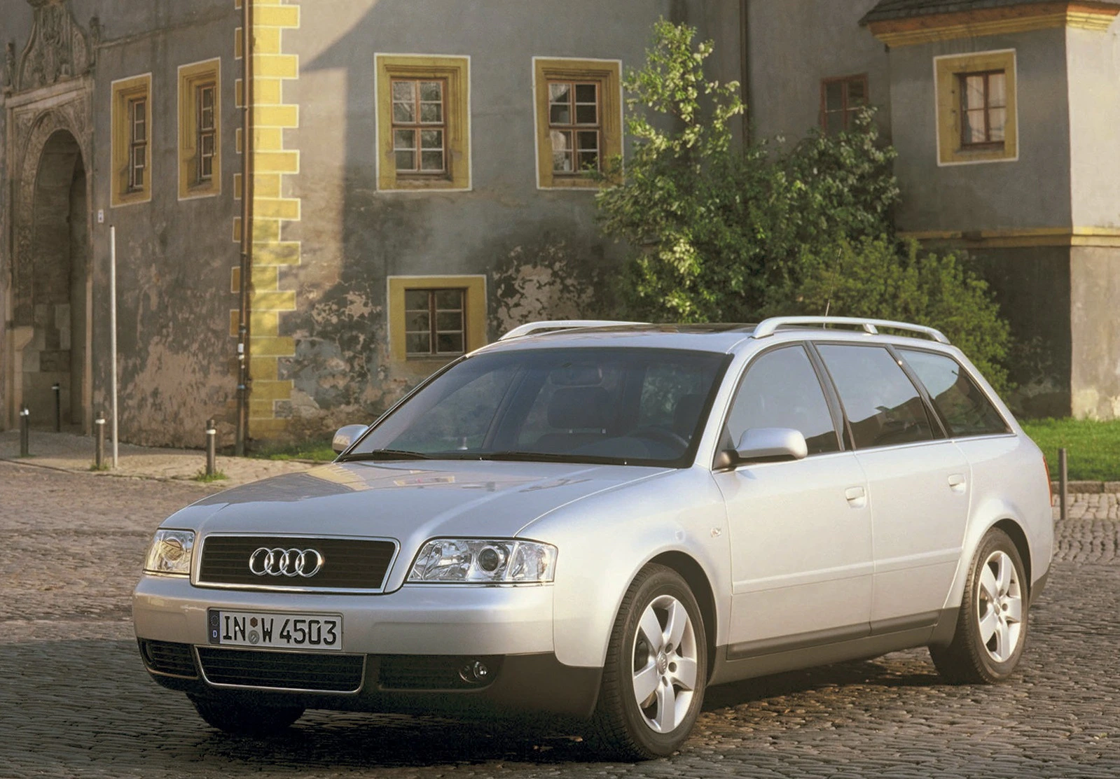 Silberner Audi A6 Avant parkt auf einem Pflastersteinplatz vor einem historischen Gebäude mit gelben Fenstern und Türen.