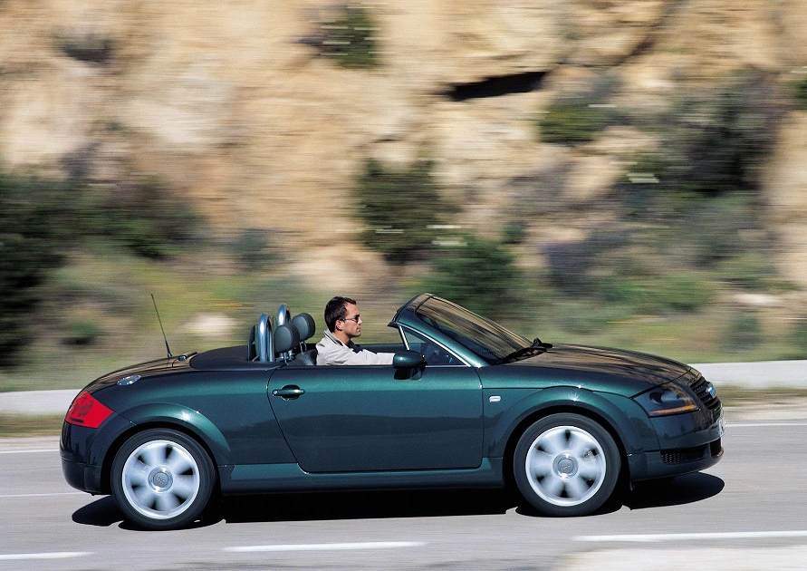 Mann fährt Cabrio auf einer Straße entlang einer felsigen Landschaft. Auto ist dunkelgrün, stilvoll und sportlich.