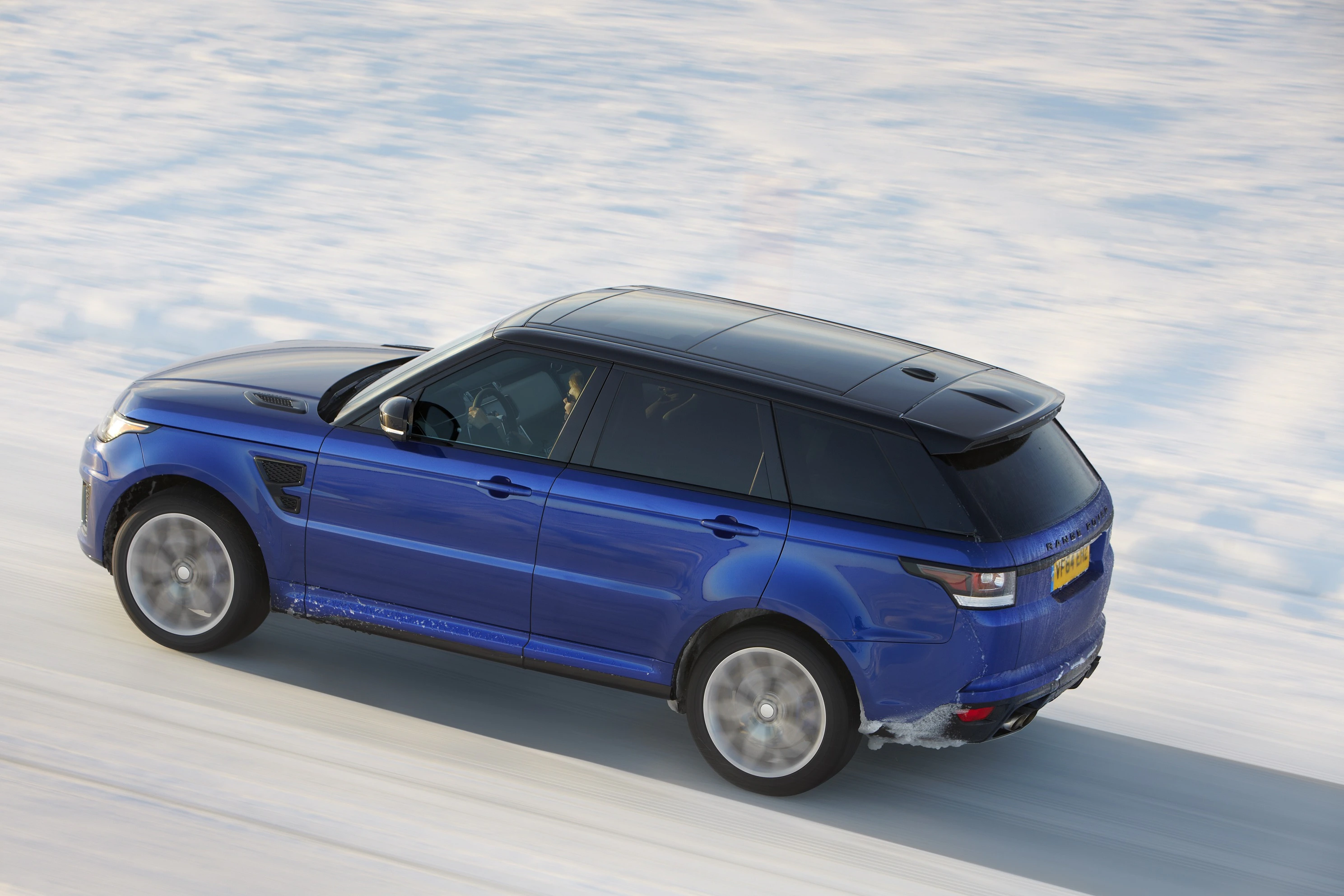 Blauer SUV fährt auf schneebedeckter Straße nach links. Der Himmel und die Felder spiegeln sich leicht auf der Karosserie.