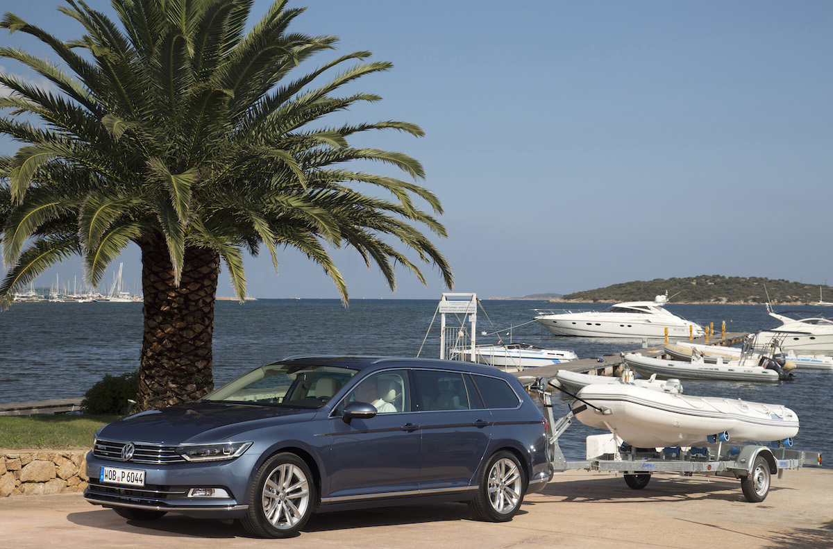 Ein blauer Kombi mit Bootsanhänger steht unter einer Palme vor einem Hafen mit mehreren Jachten und Segelbooten.