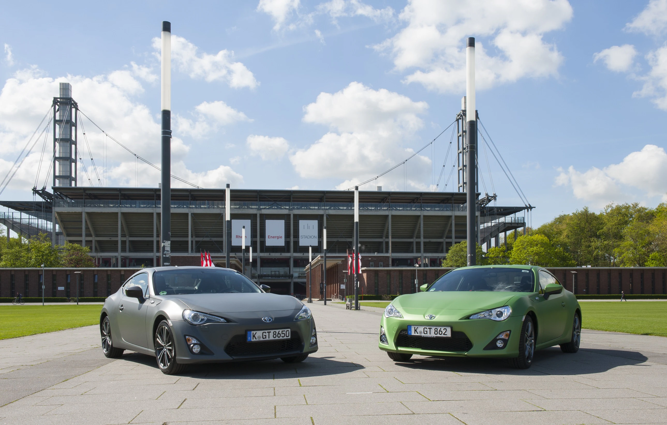 Zwei Sportwagen stehen nebeneinander vor einem Stadion bei sonnigem Wetter. Links ist ein grauer, rechts ein grüner Wagen.