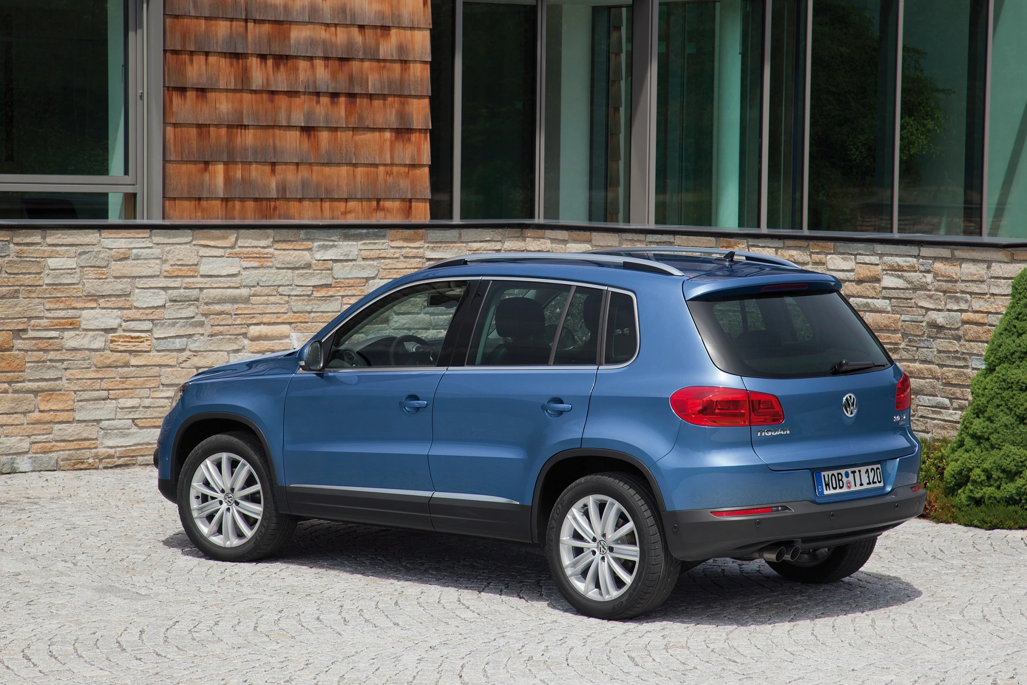 Blauer SUV, geparkt auf gepflastertem Hof vor einem modernen Gebäude mit Stein- und Holzwänden.