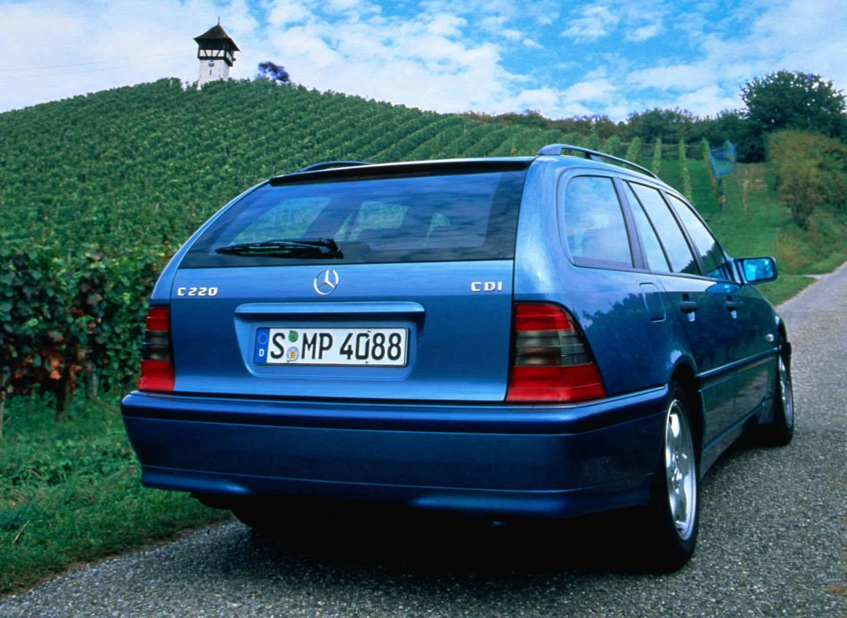 Blauer Mercedes C220 Kombi auf einer Straße, rechts Weinberge und ein kleiner Turm im Hintergrund. Wolkiger Himmel.