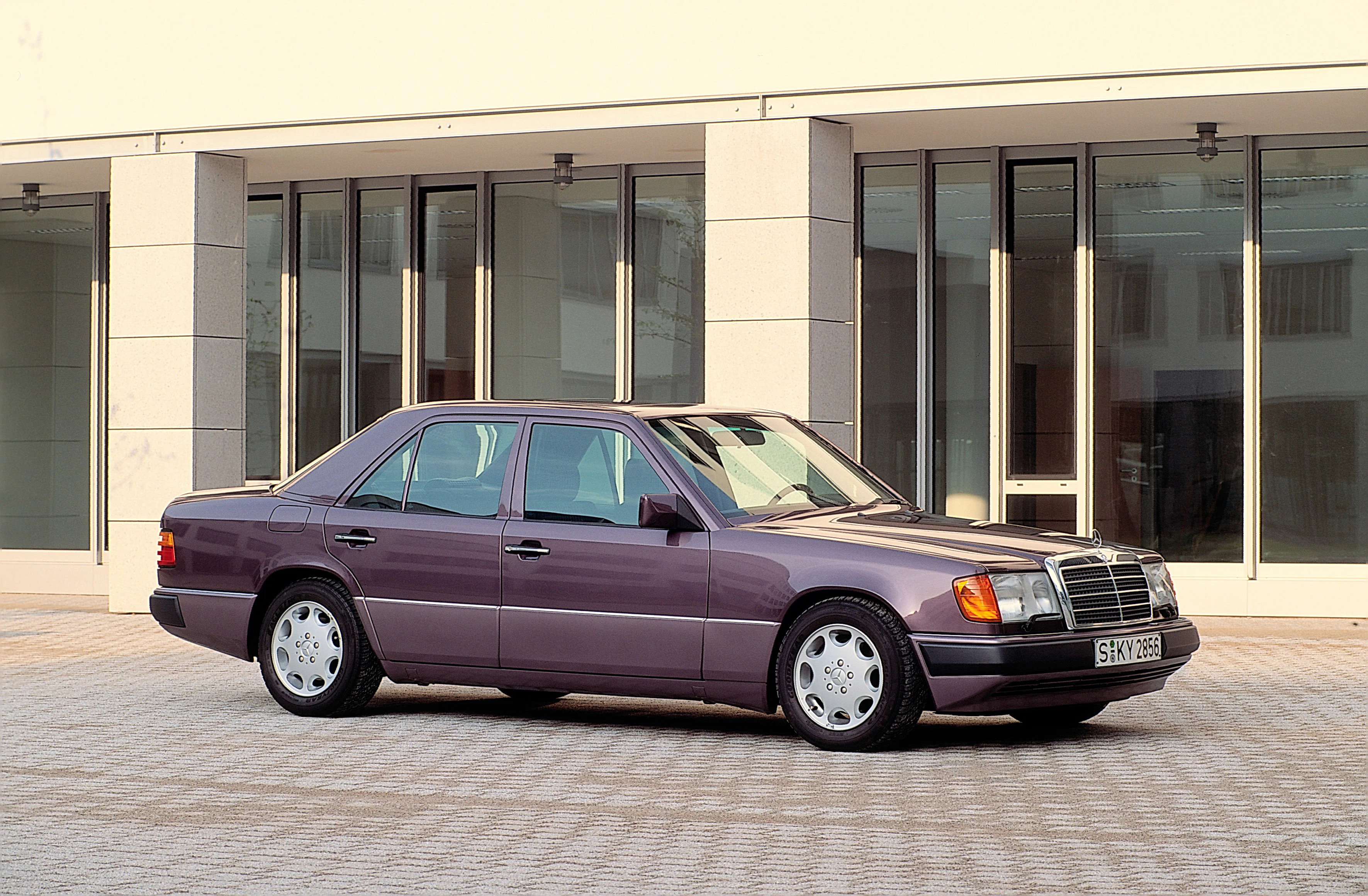 Lila Mercedes-Benz W124 Limousine, seitlich abgebildet, steht vor einem modernen Gebäude mit großen Fenstern.