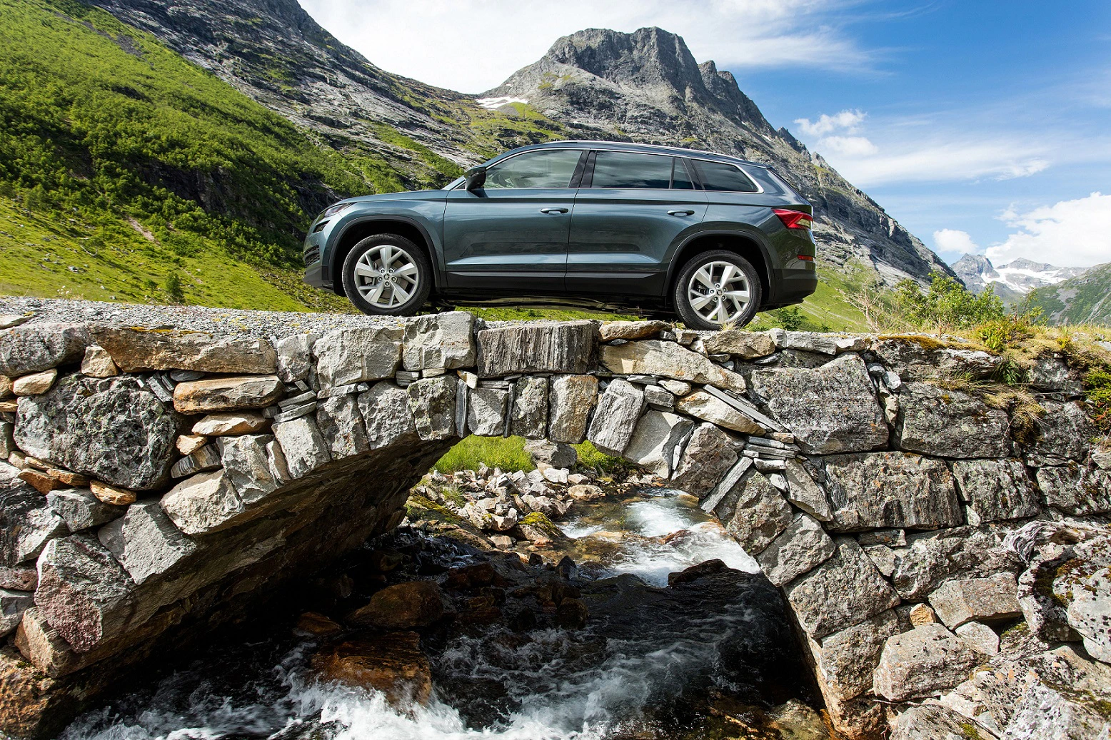 Ein SUV fährt über eine alte Steinbrücke in einer bergigen Landschaft mit blauem Himmel und einem fließenden Fluss.