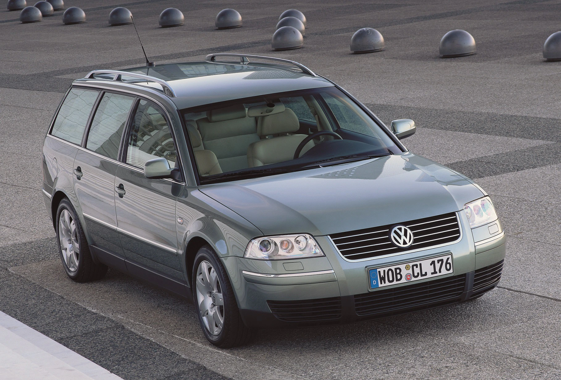 Silberner Volkswagen Passat Variant der frühen 2000er auf gepflasterter Fläche, städtische Umgebung, futuristische Architektur im Hintergrund.