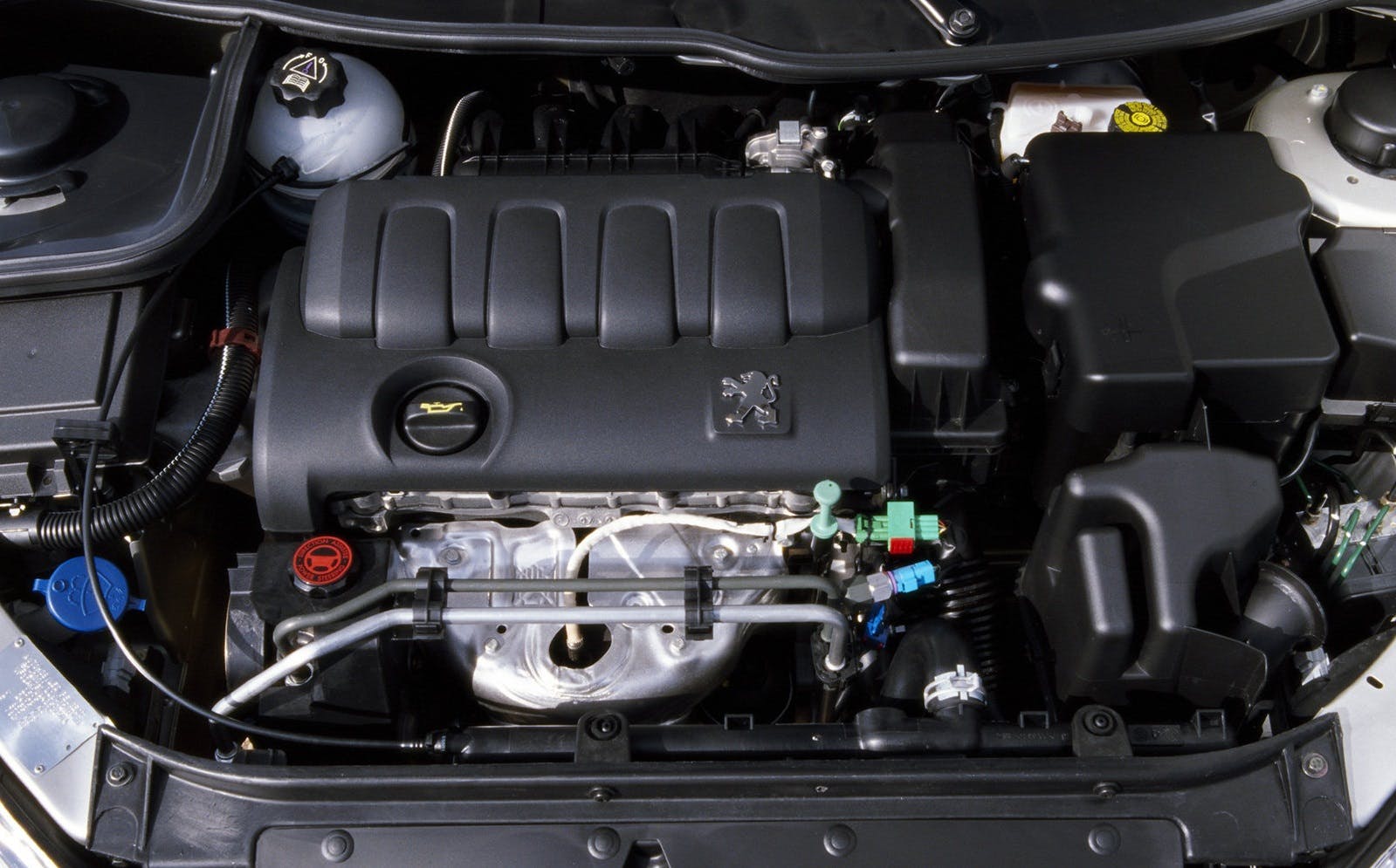 Motorraum eines Autos mit sichtbarem Motorblock, Kabeln und Komponenten. Schwarze Abdeckung mit einem Markenemblem in der Mitte.