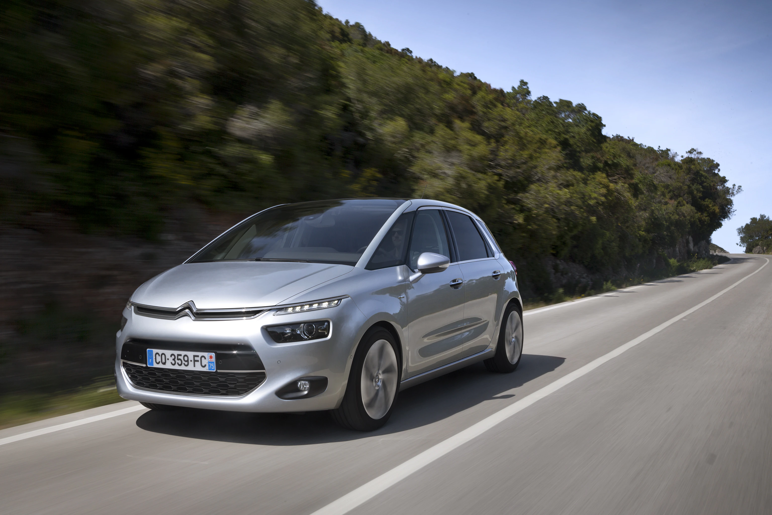 Silberner Citroën C4 Picasso fährt auf einer Landstraße entlang einer grünen, bewaldeten Gegend an einem sonnigen Tag.