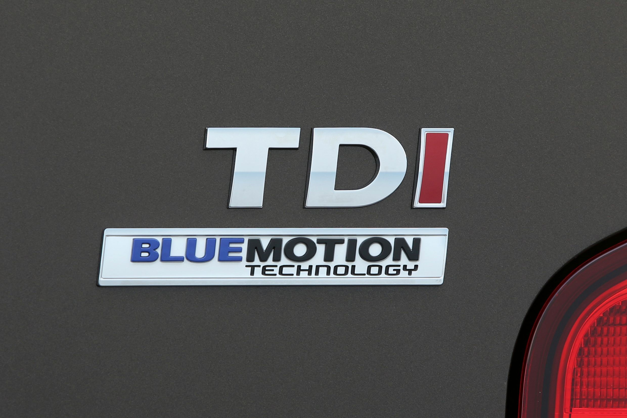 Nahaufnahme eines Autos mit "TDI" und "BlueMotion Technology"-Logo auf dunklem Hintergrund. Teil eines Rücklichts ist sichtbar.