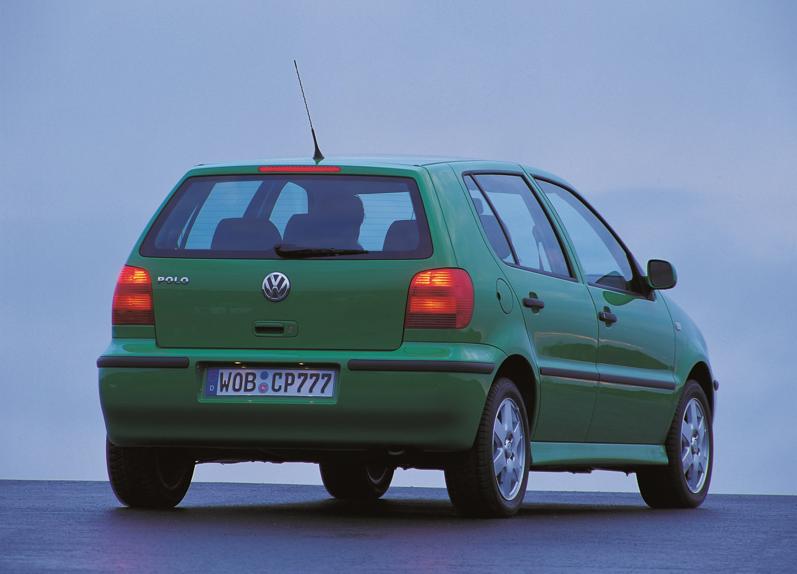 Grüner VW Polo der 1990er Jahre steht draußen auf einer Straße, fotografiert von hinten vor einem bewölkten Himmel.