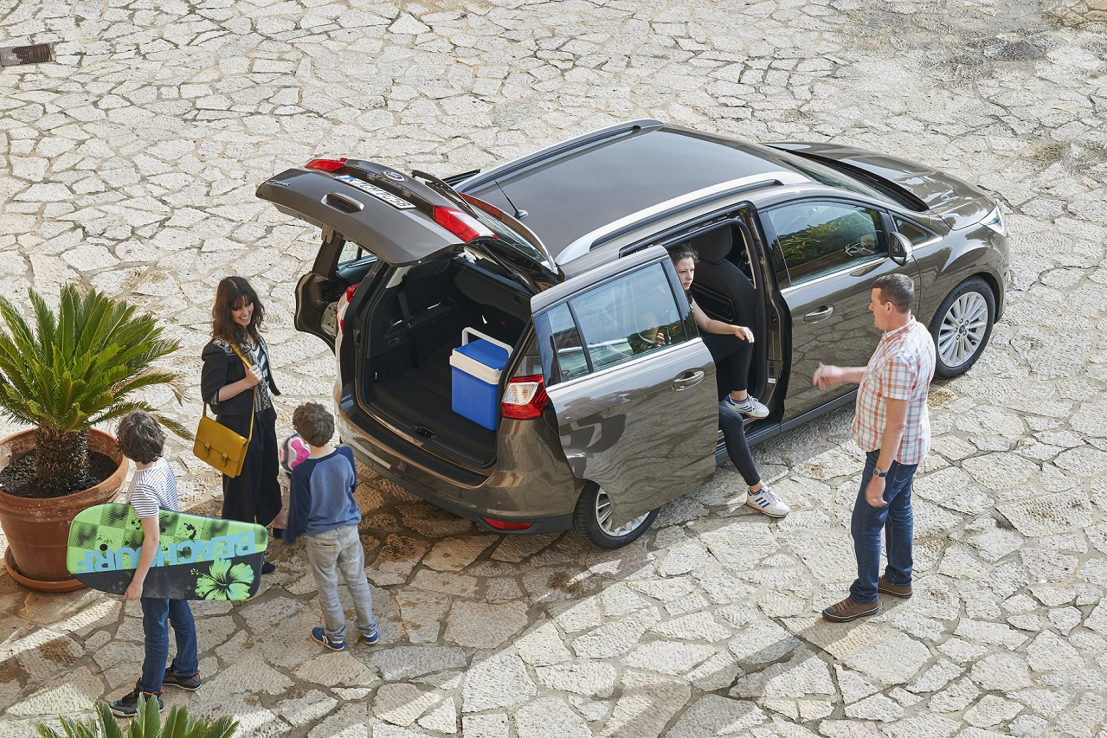 Eine Familie steht neben einem Auto auf einem Steinboden. Ein Kind hält ein Skateboard, während ein Elternteil einen Kühler im Kofferraum überprüft.