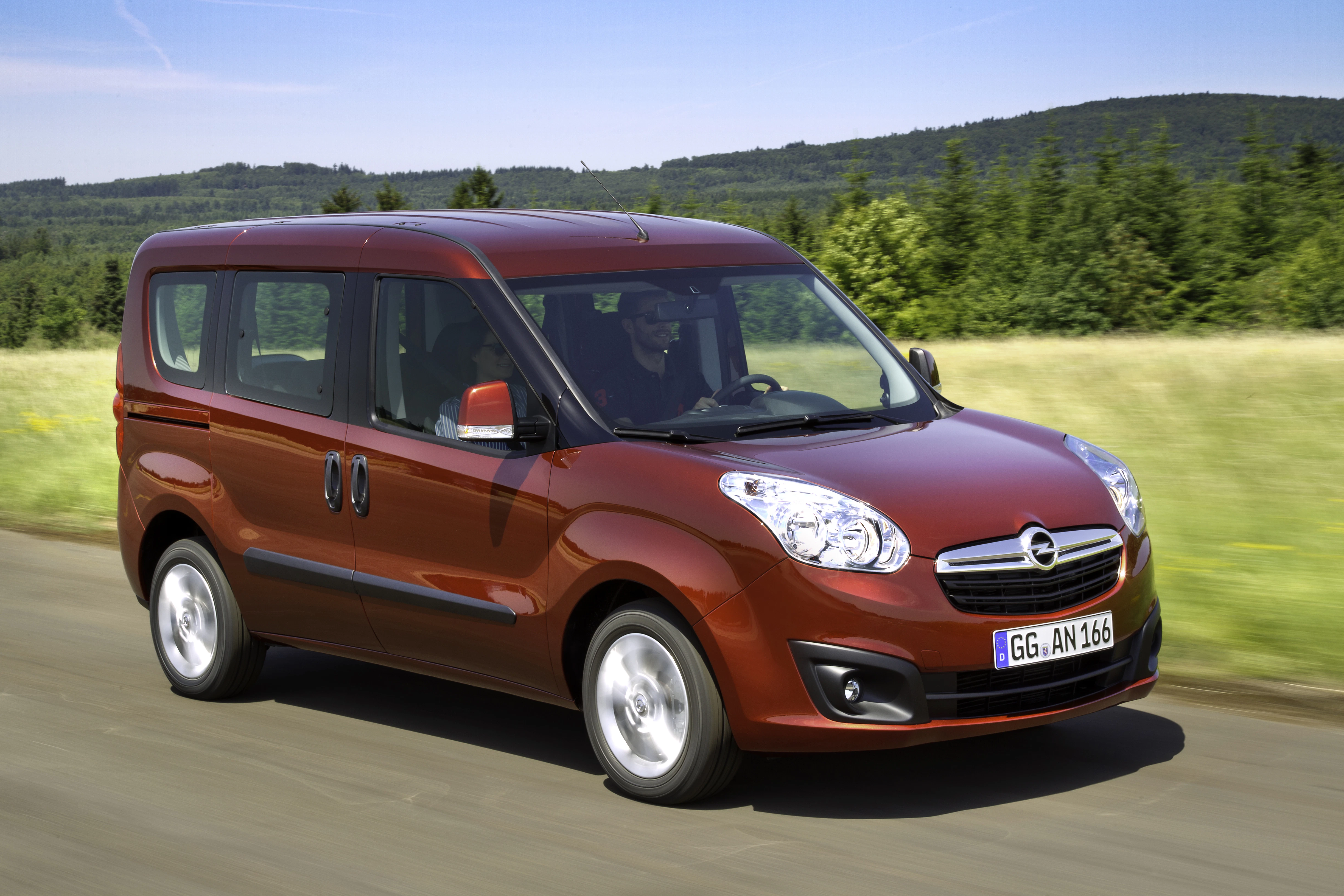 Roter Opel-Van fährt auf einer Landstraße durch eine grüne Landschaft mit Wald im Hintergrund.