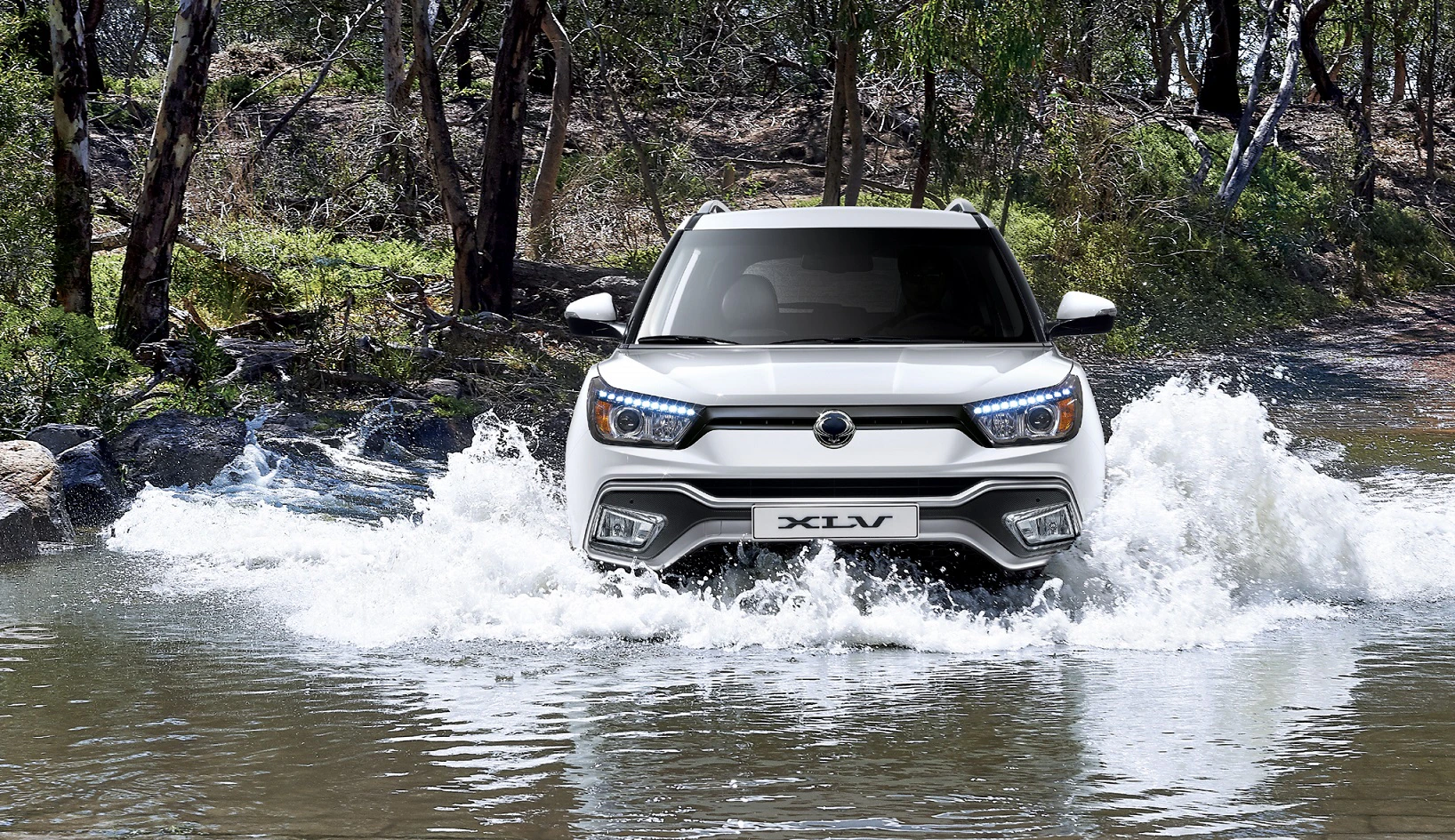 Ein weißer SUV fährt durch seichtes Wasser in einer bewaldeten Umgebung.