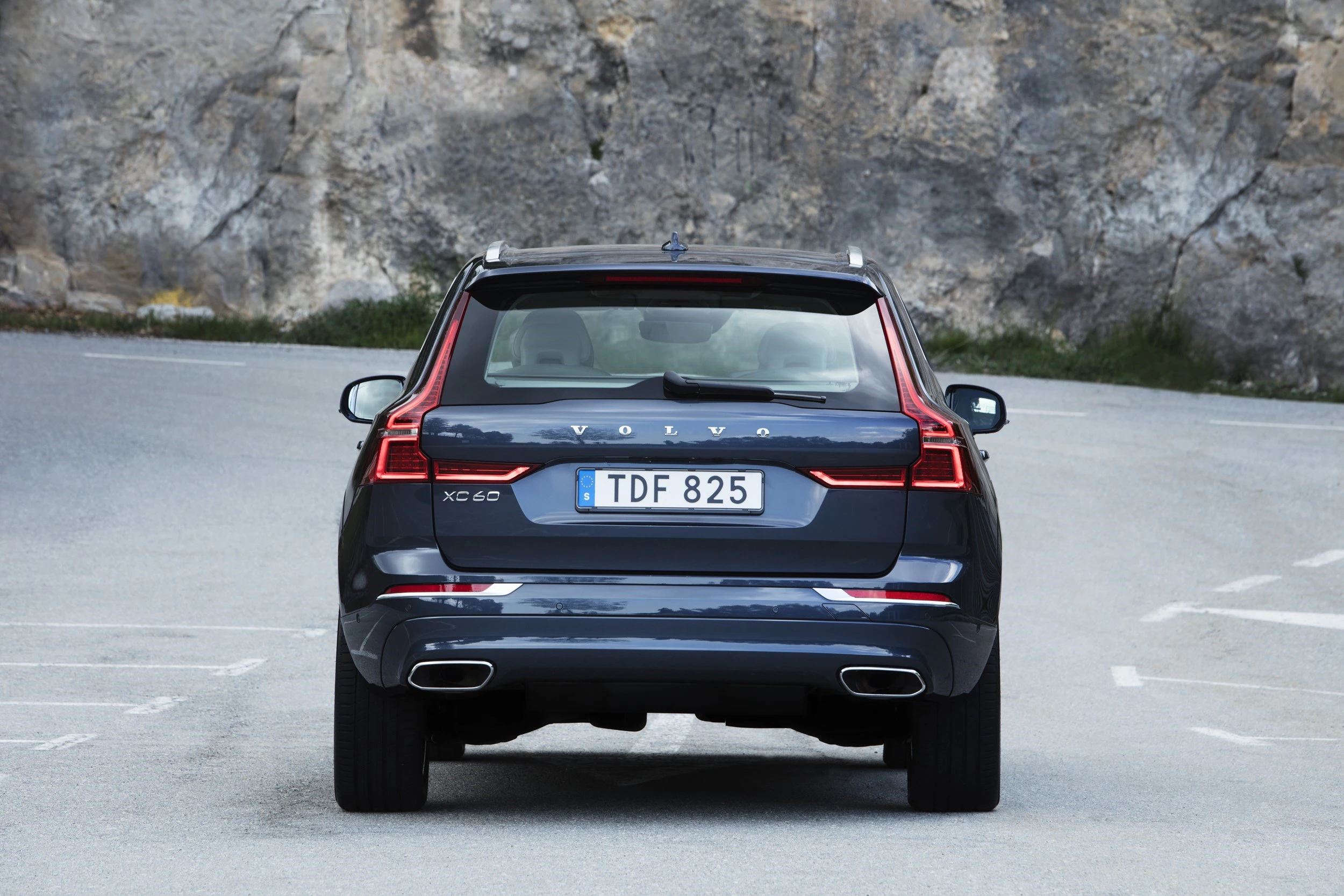Rückansicht eines dunkelblauen Volvo XC60 auf einer Straße vor einer Felswand, mit beleuchteten Rücklichtern und einem Kennzeichen.