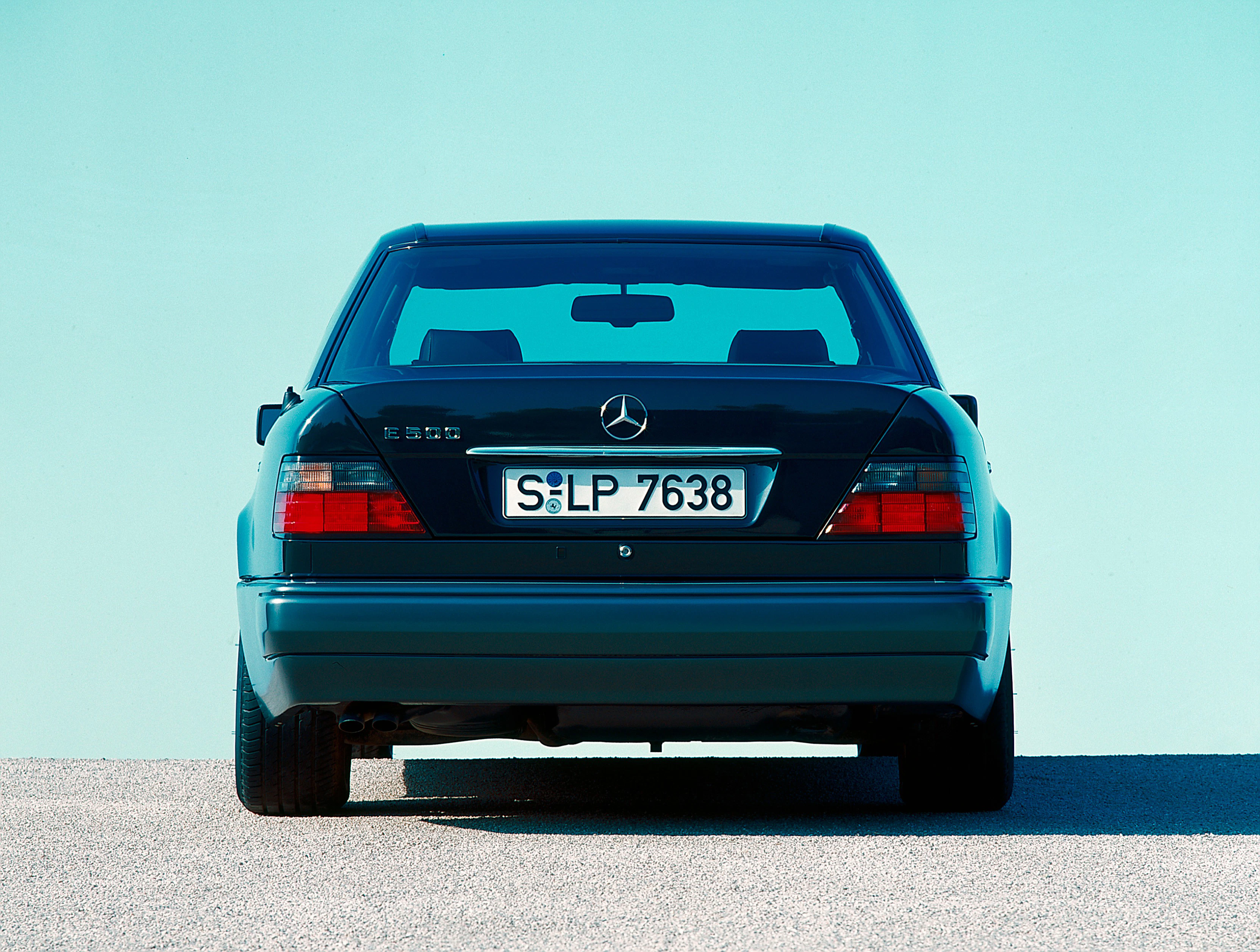 Rückansicht eines schwarzen Mercedes-Benz E500 vor einer turkisen Wand, Nummernschild "S-LP 7638", auf einer asphaltierten Fläche.