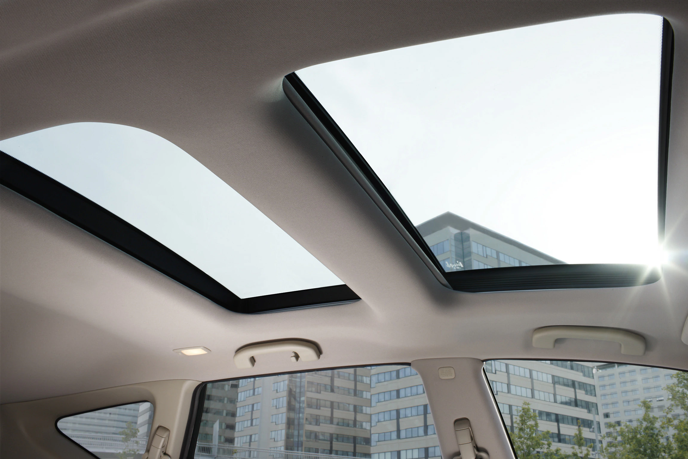 Innenansicht eines Autos mit zwei offenen, rechteckigen Schiebedächern, darunter moderne Bürogebäude und Sonnenlicht im Hintergrund.