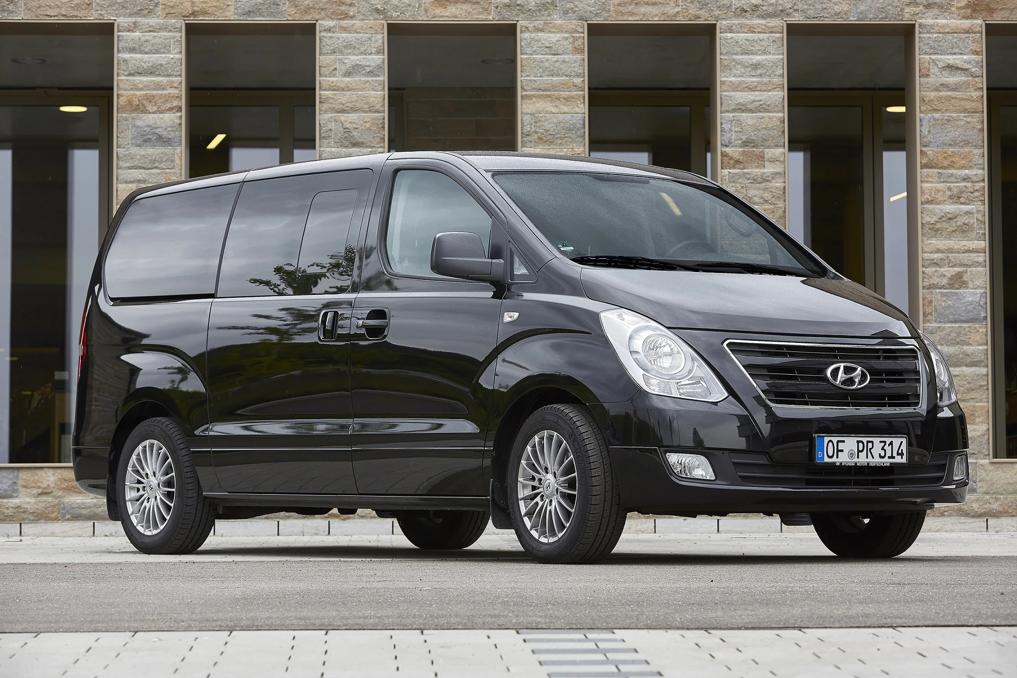 Schwarzer Hyundai Kleinbus parkt vor einem Gebäude mit Steinmauer und großen Fenstern, seitlich von der Kamera aufgenommen.