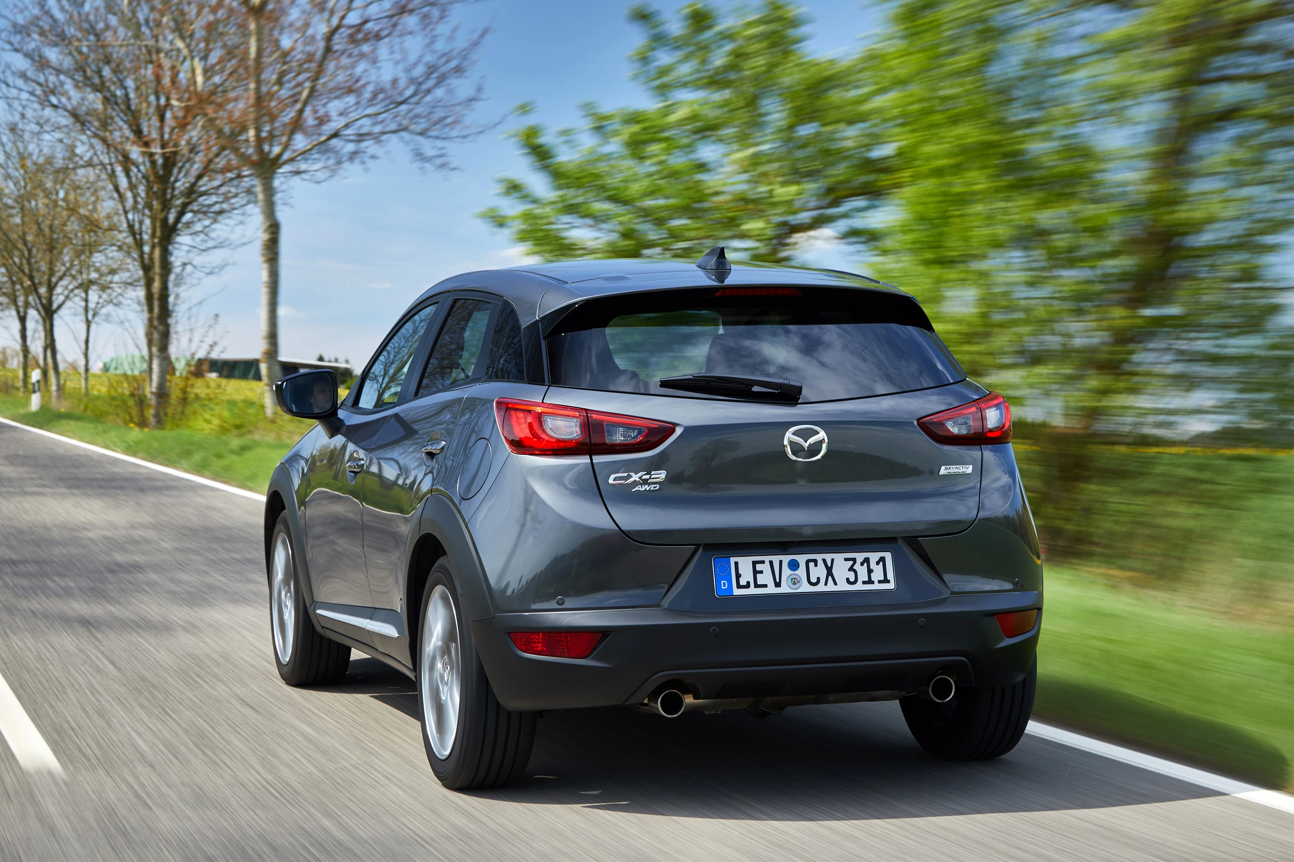 Grauer Mazda CX-3 fährt auf einer Landstraße, umgeben von Bäumen und Wiesen.