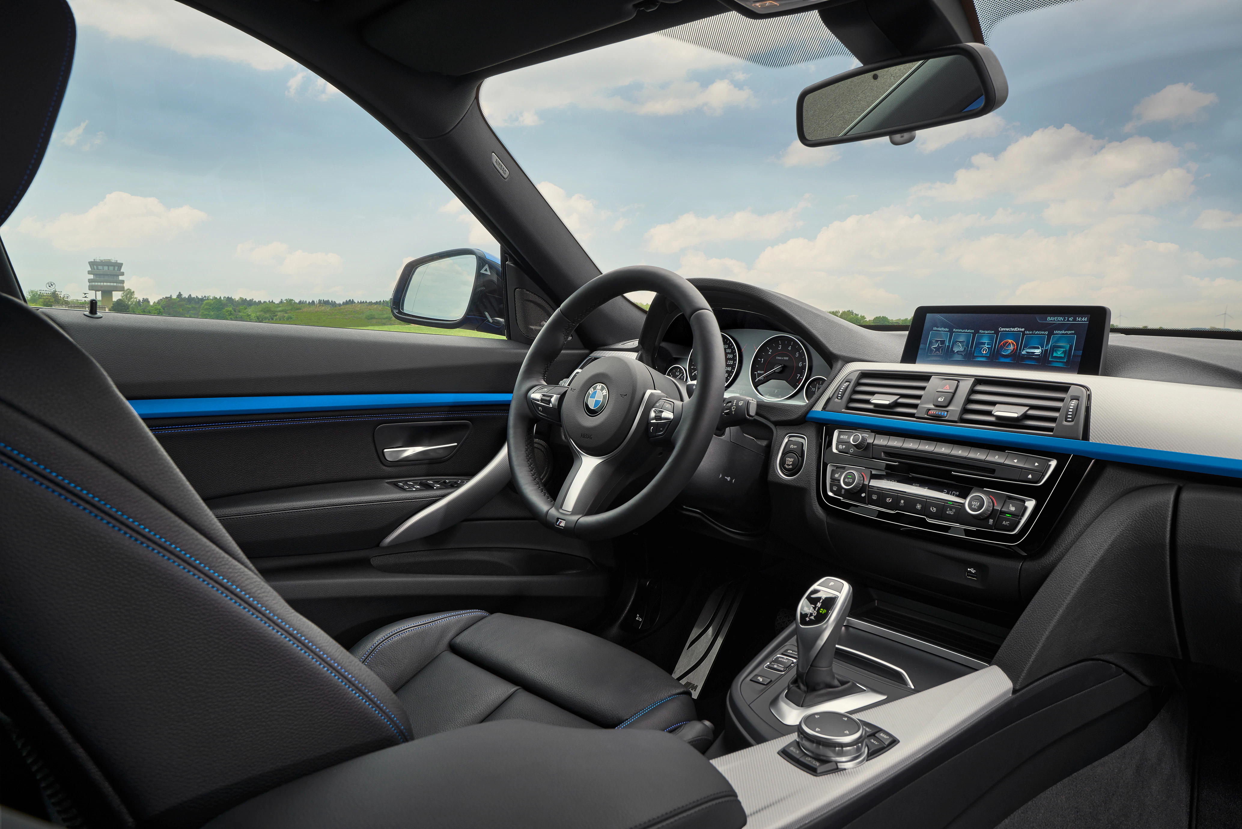 Innenraum eines modernen BMW mit schwarzer Lederpolsterung, Multifunktionslenkrad, Navigationssystem und großem Touchscreen bei bewölktem Himmel.