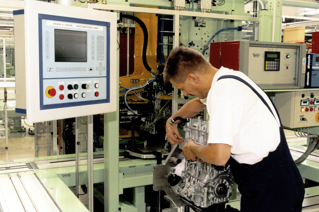 Ein Mann in Arbeitskleidung montiert einen Motor in einer Fabrik, umgeben von Maschinen und einem Steuerpanel links im Bild.