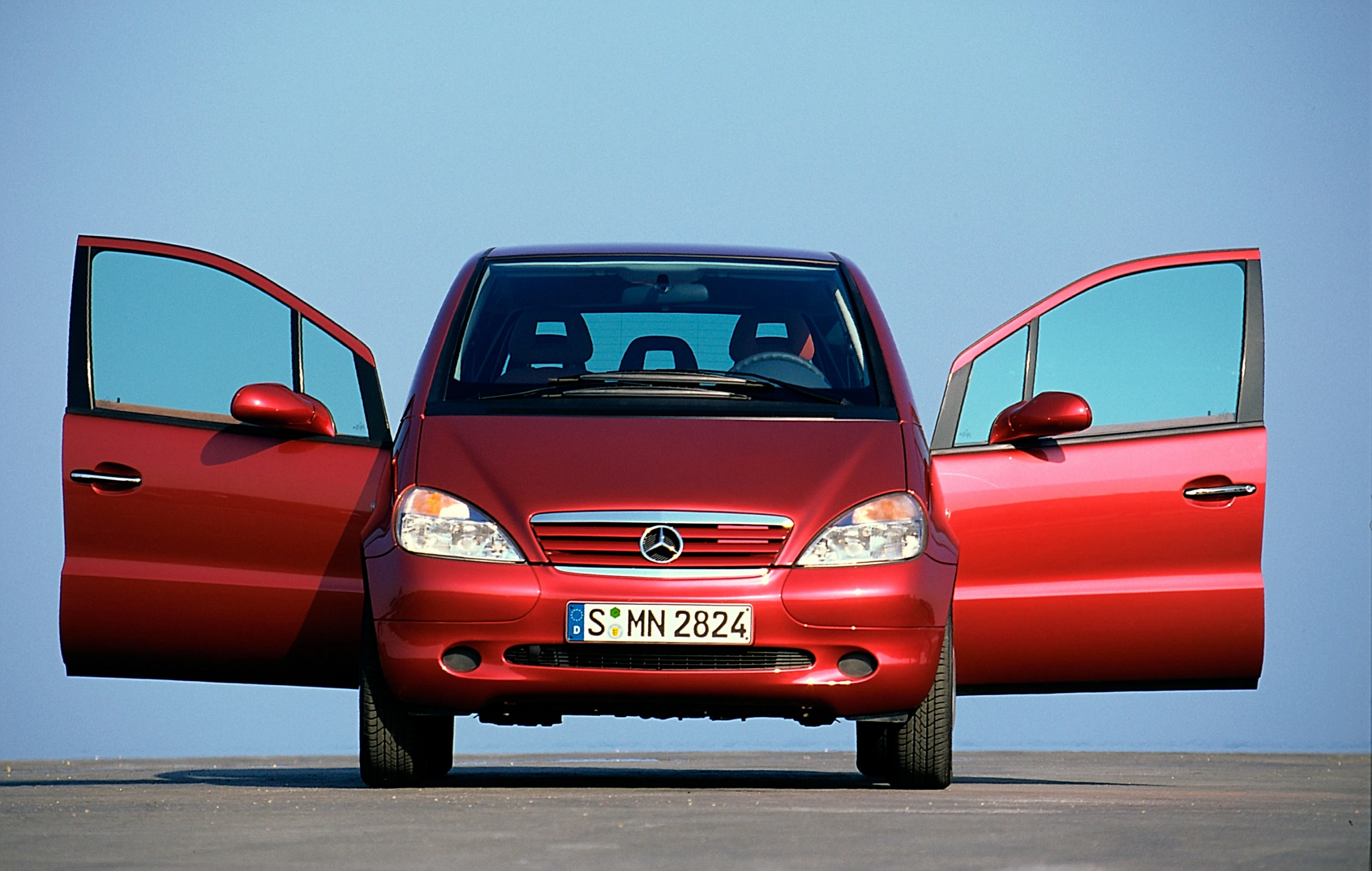 Rotes Auto mit geöffneten vorderen Seitentüren, frontal auf einer Straße stehend, klarer blauer Himmel im Hintergrund.