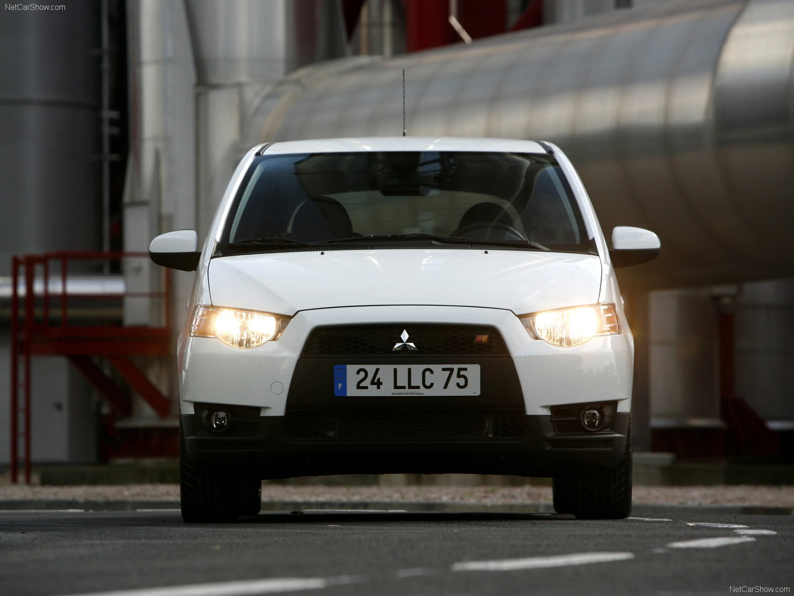 Weißer Mitsubishi Lancer von vorne, geparkt vor Industrieanlagen mit großen Metallrohren. Die Scheinwerfer sind eingeschaltet.