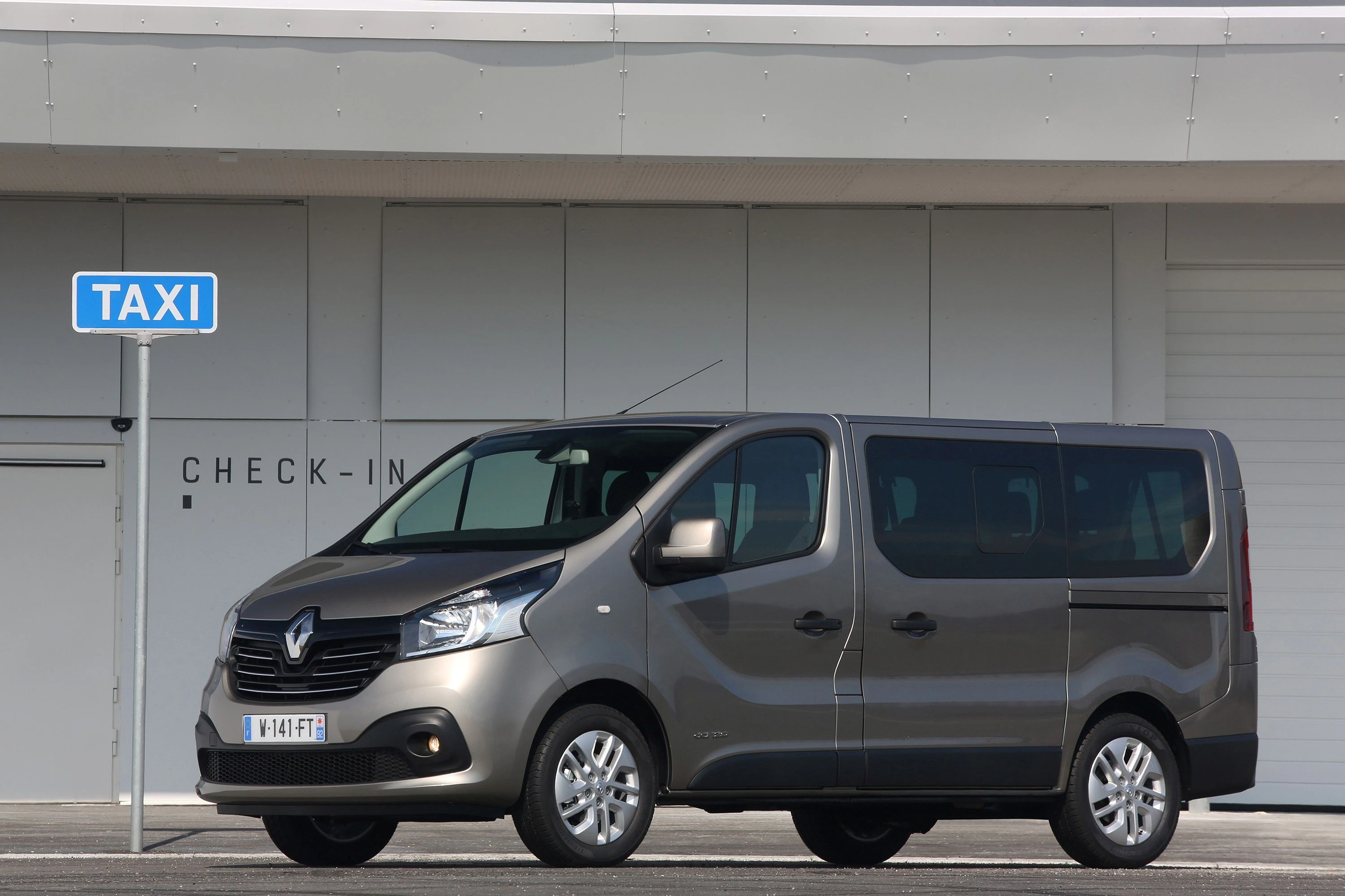 Grauer Renault Van als Taxi vor einem Gebäude geparkt, neben einem blauen TAXI-Schild.