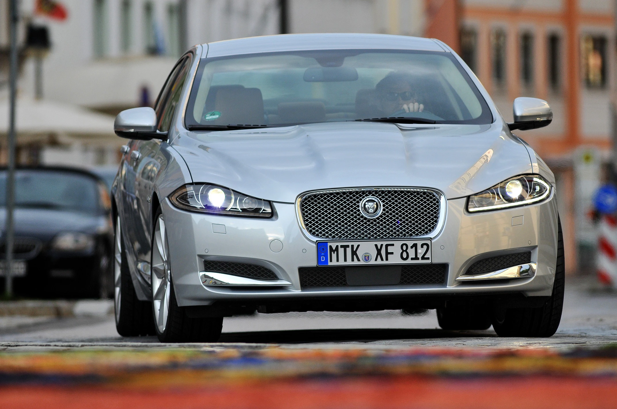 Silberner Jaguar XF fährt auf bunter Straße in Stadtumgebung. Deutsche Nummernschilder, modernes Design und markantes Kühlergrill.