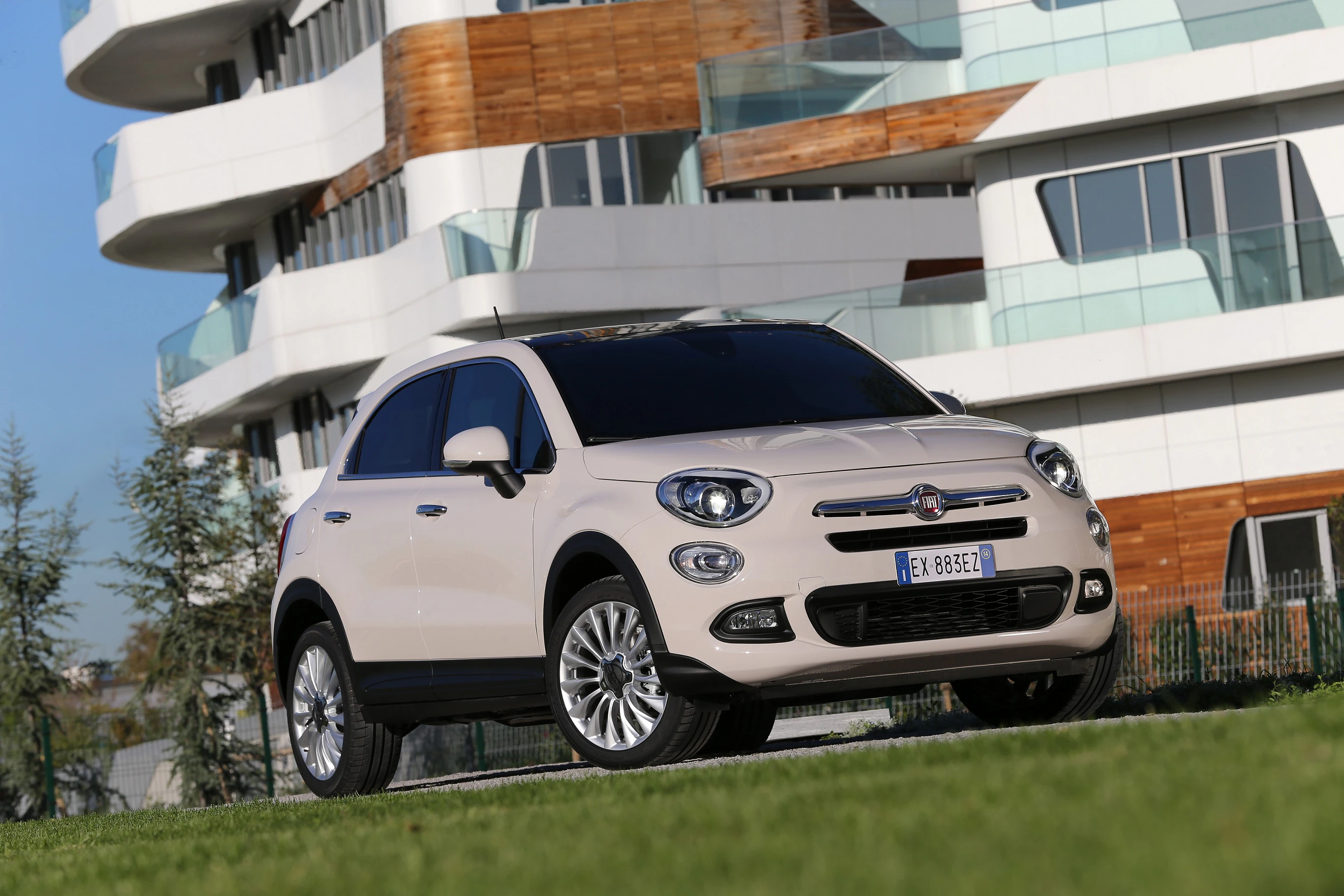 Beiger SUV parkt vor modernem Gebäude mit Glasbalkonen und Holzdetails, blauer Himmel im Hintergrund.