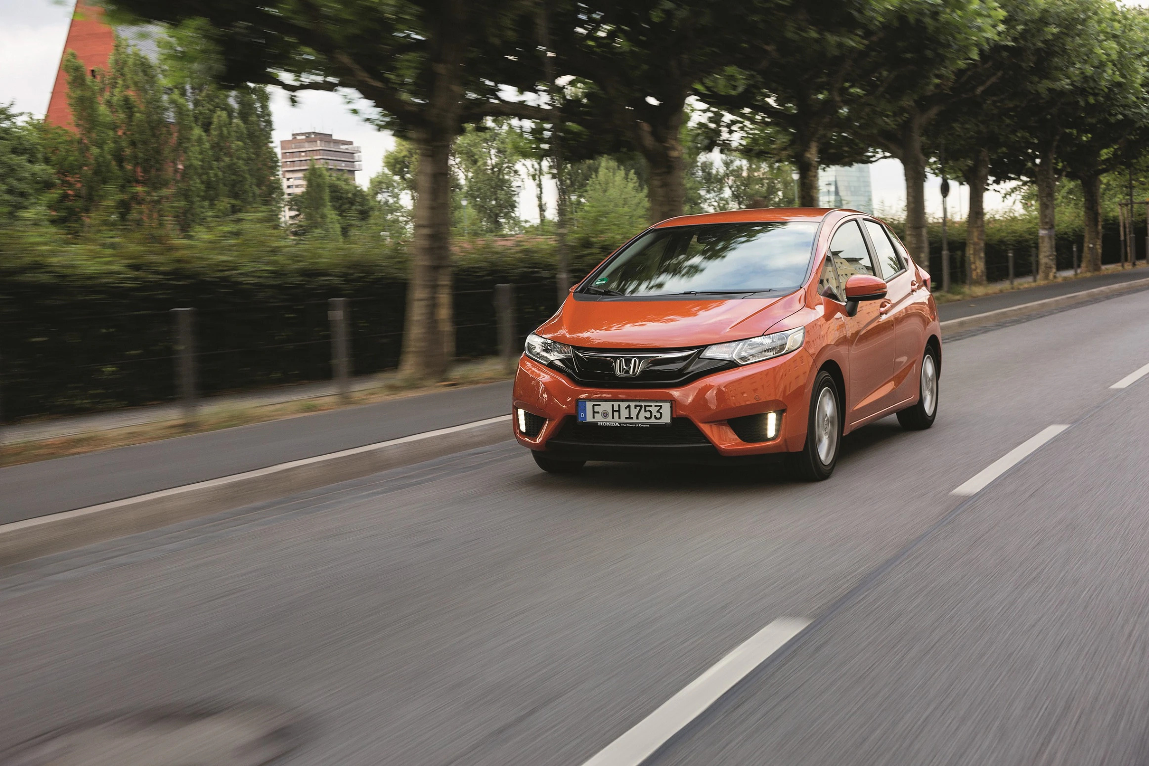 Orangefarbener Honda Kleinwagen fährt auf einer städtischen Straße, umgeben von Bäumen.