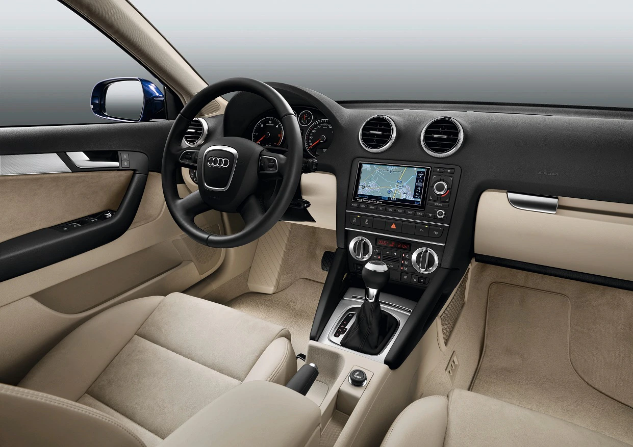 Innenraum eines Autos mit beigem Interieur, Lenkrad mit Audi-Logo, Navigationssystem auf der Mittelkonsole und Schaltknüppel.