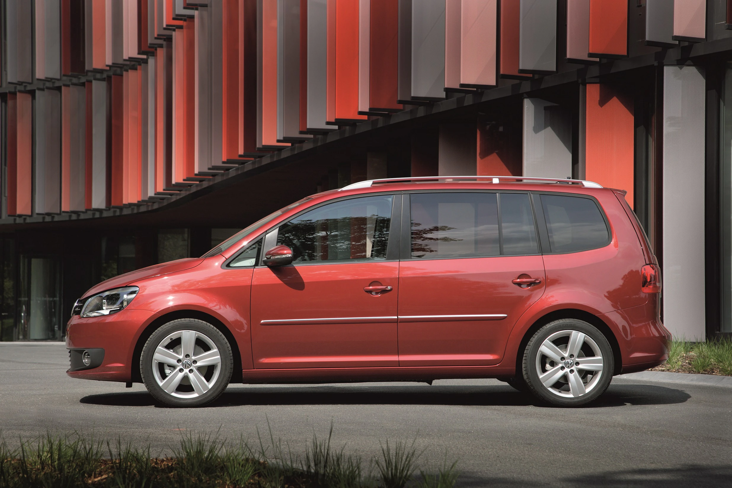 Roter Minivan parkt vor einem modernen Gebäude mit rot-schwarzer Fassade. Fahrzeug hat silberne Felgen und große Fenster.