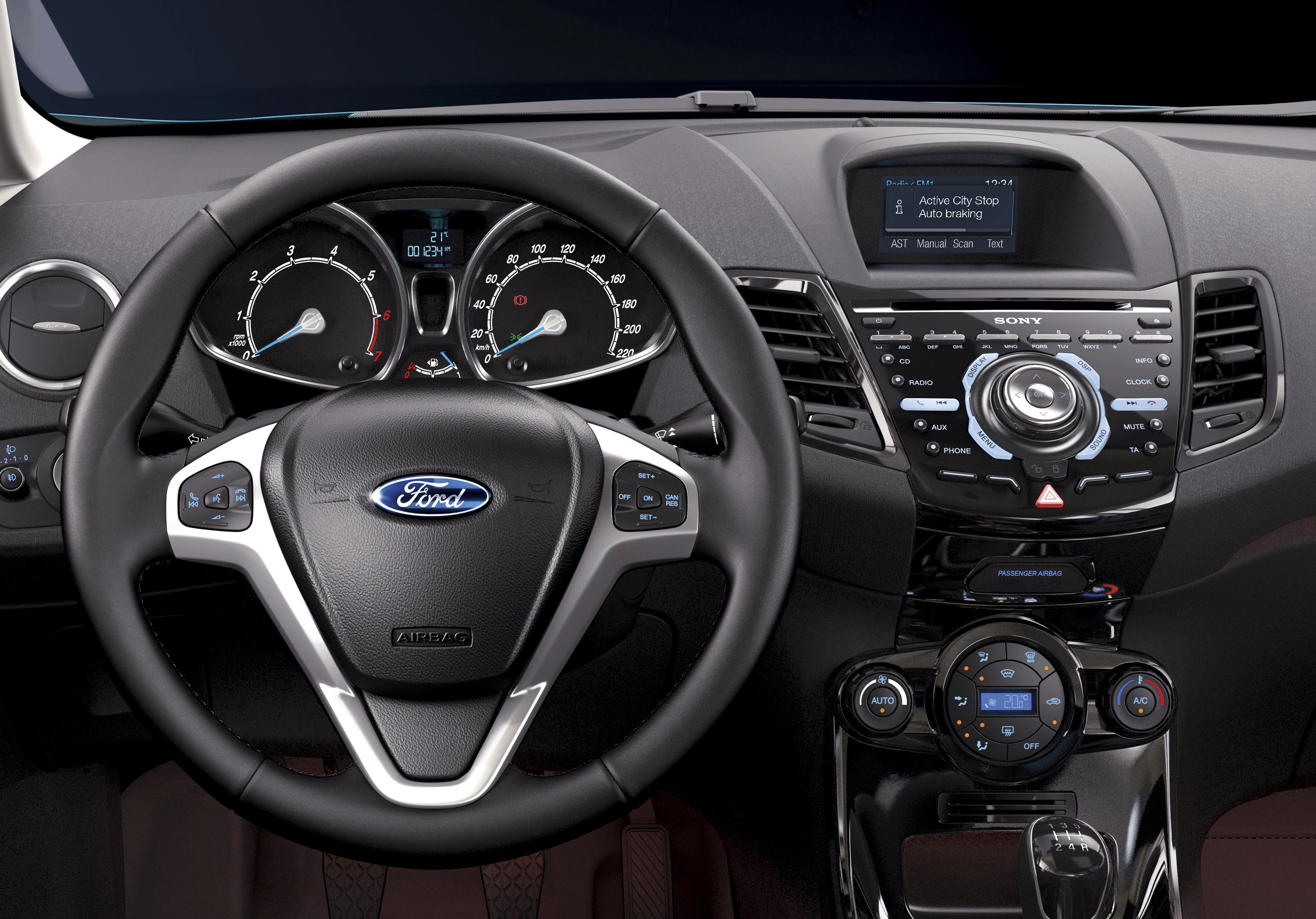Nahaufnahme eines Fahrzeug-Cockpits mit Ford-Logo, Lenkrad, Tachometer und Mittelkonsole mit Bildschirm und Steuerknöpfen.