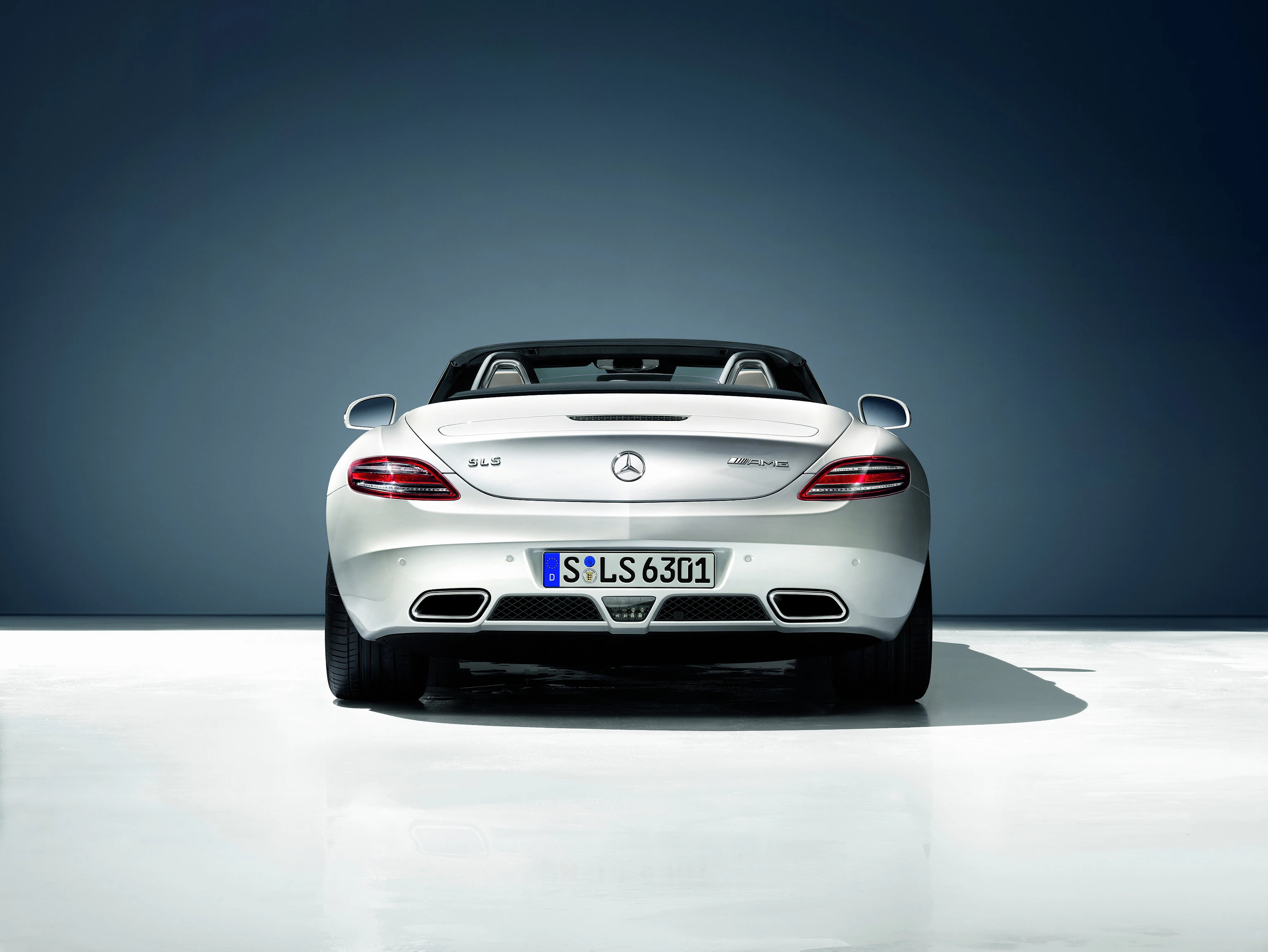 Silberner Mercedes-Benz SLS AMG Roadster von hinten auf weißem Boden vor dunklem Hintergrund, Nummernschild sichtbar.
