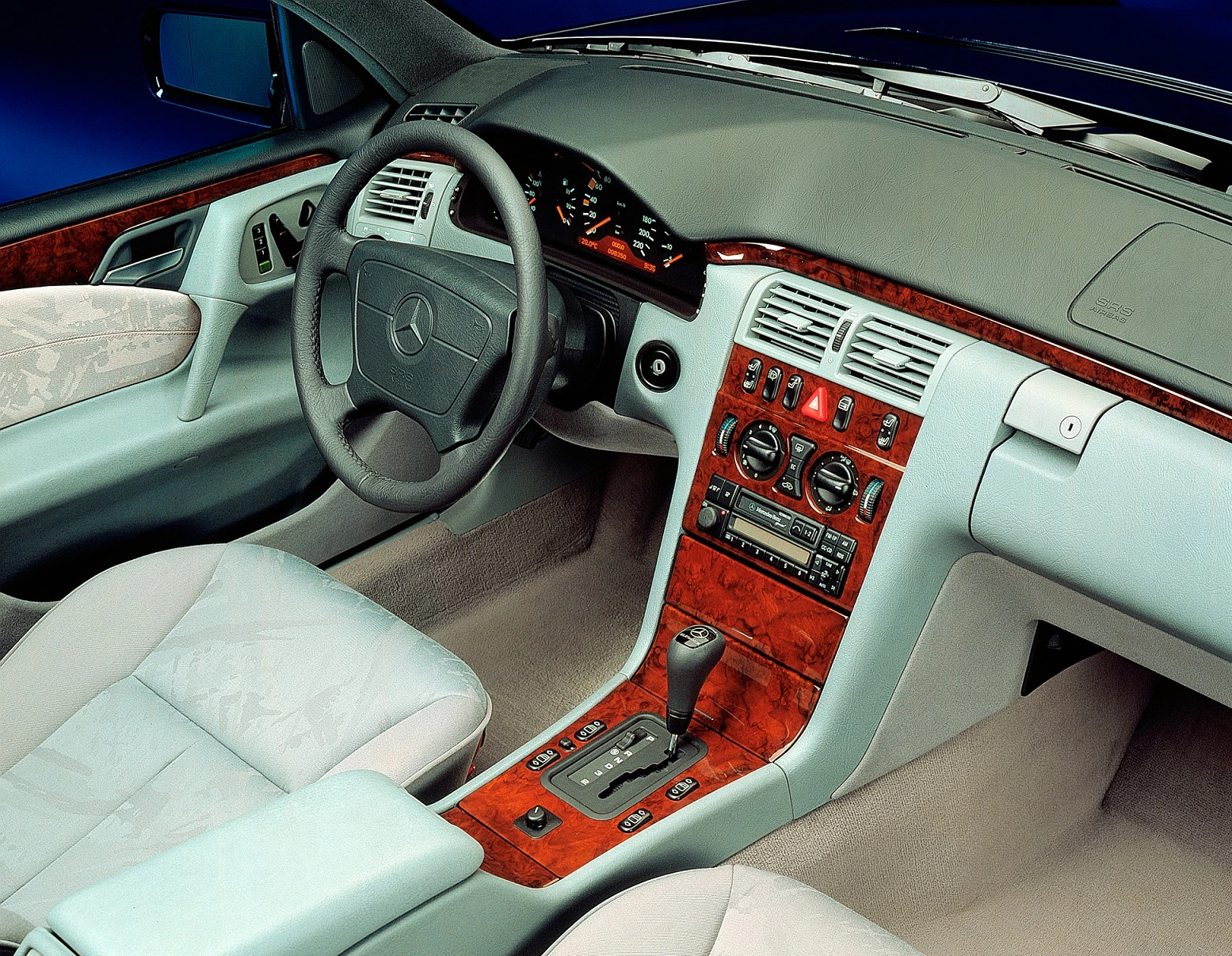 Innenraum eines Mercedes-Benz mit hellem Ledersitz, Holzverkleidung, Automatikgetriebe und Lenkrad mit prominentem Logo.