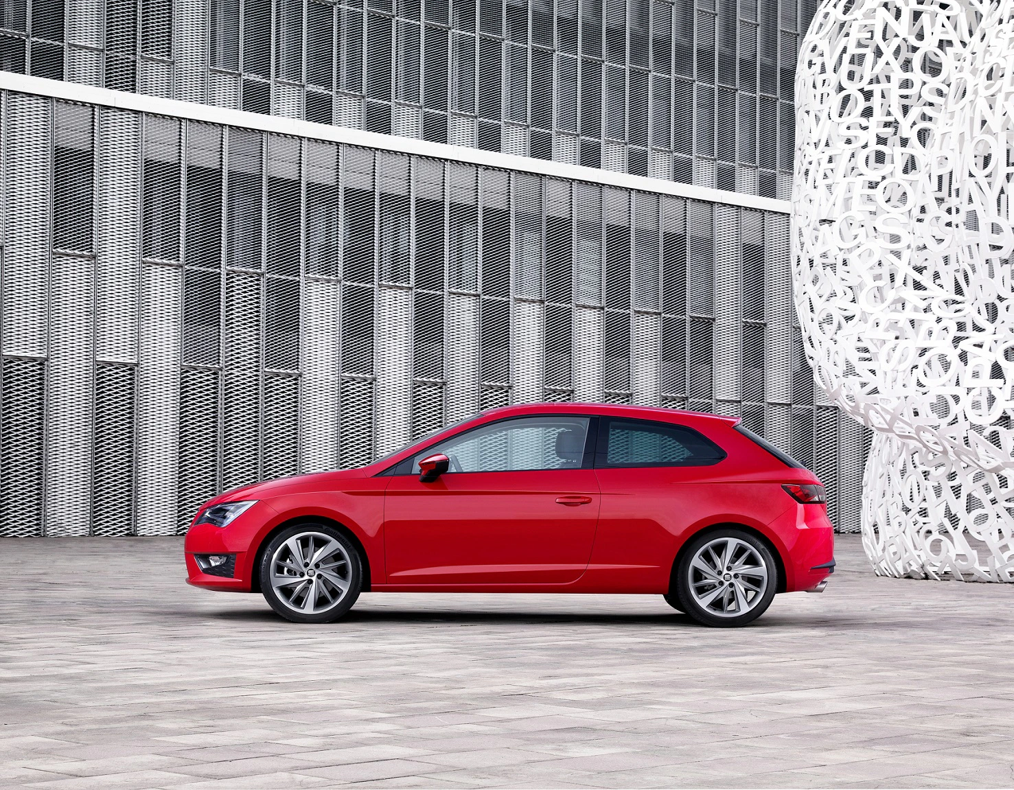Roter Sportwagen vor moderner Gebäudefassade und kunstvoller Skulptur. Urbaner Hintergrund mit geometrischem Design.