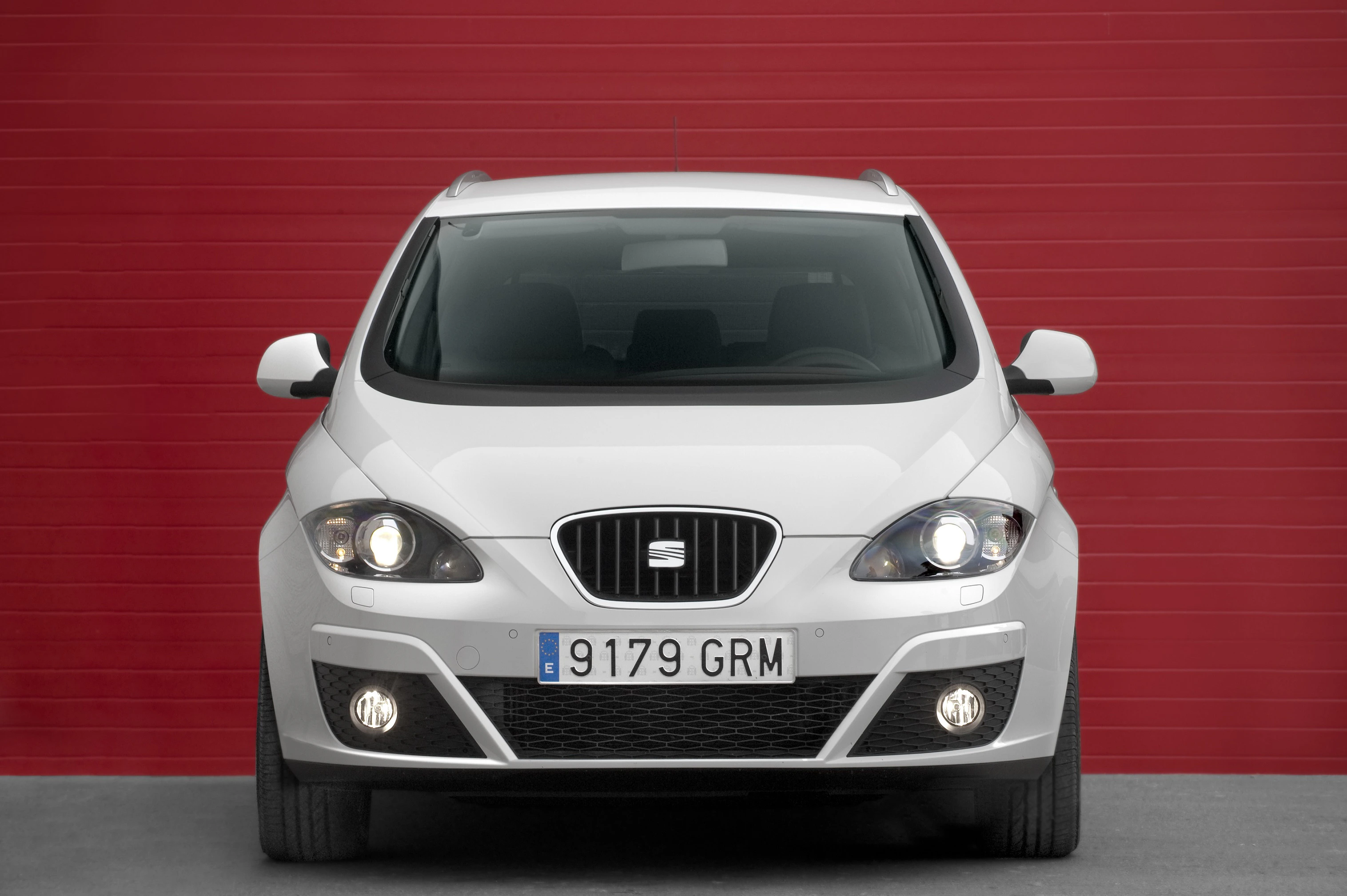 Weißer SEAT Altea steht vor einer roten, horizontal gerippten Wand. Fahrzeug von vorne fotografiert, Scheinwerfer und Kühlergrill sichtbar.