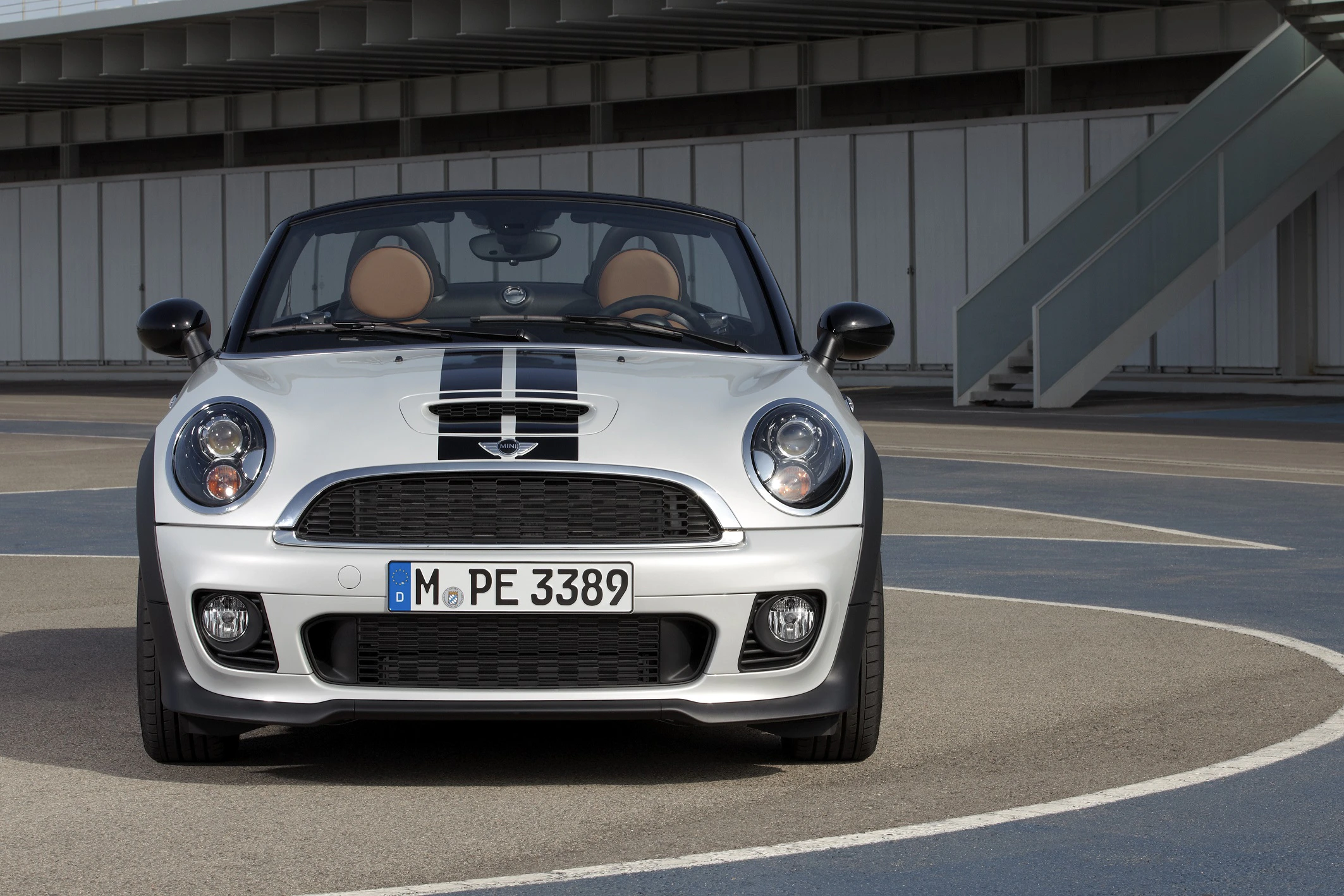Silberner Mini Cooper Cabrio mit schwarzen Streifen, fotografiert von vorne auf einem offenen Parkplatz vor einem modernen Gebäude.