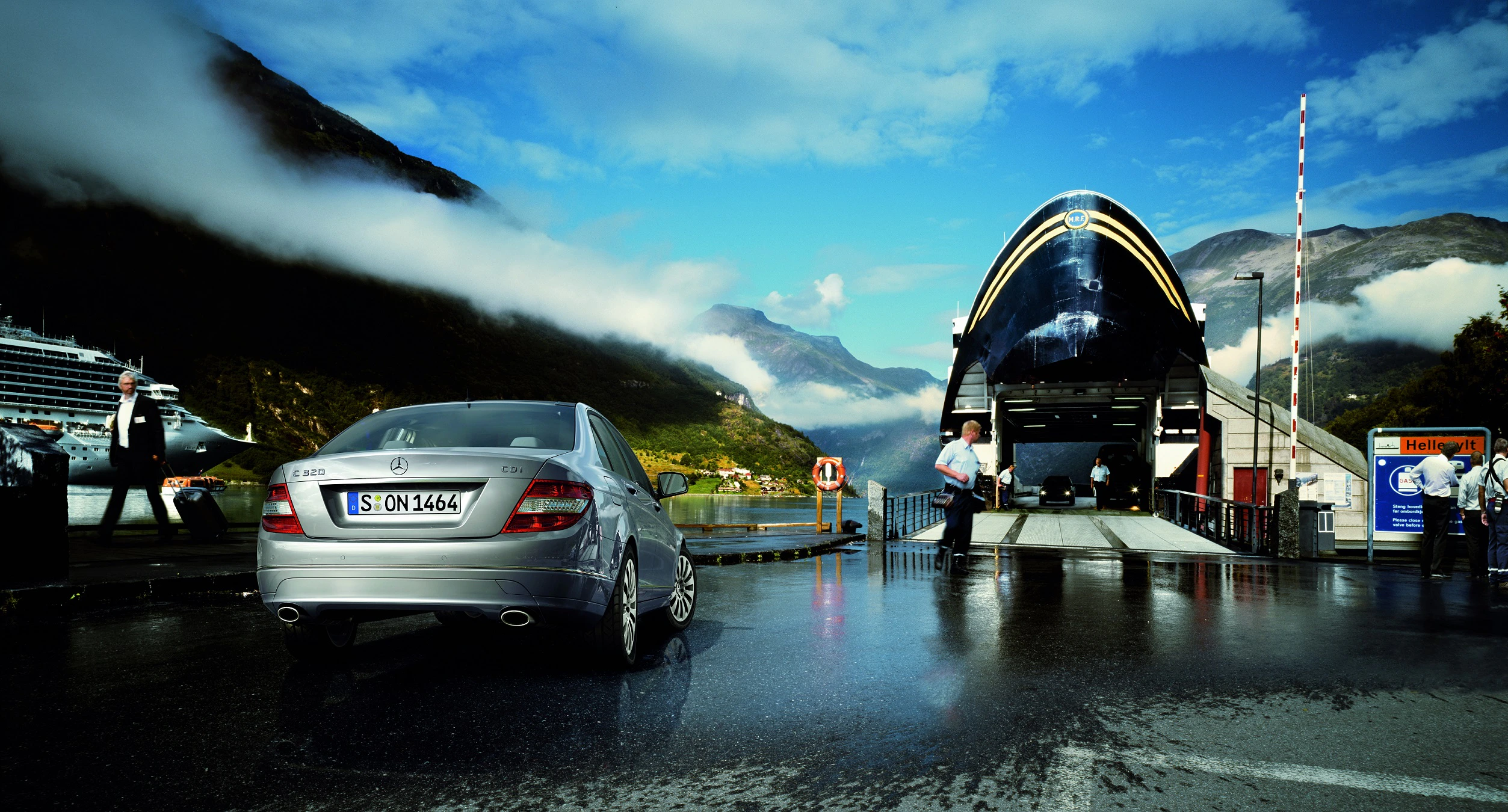 Ein silbernes Auto wartet vor einer Fähre in einer malerischen norwegischen Fjordlandschaft mit Bergen und Wolken im Hintergrund.