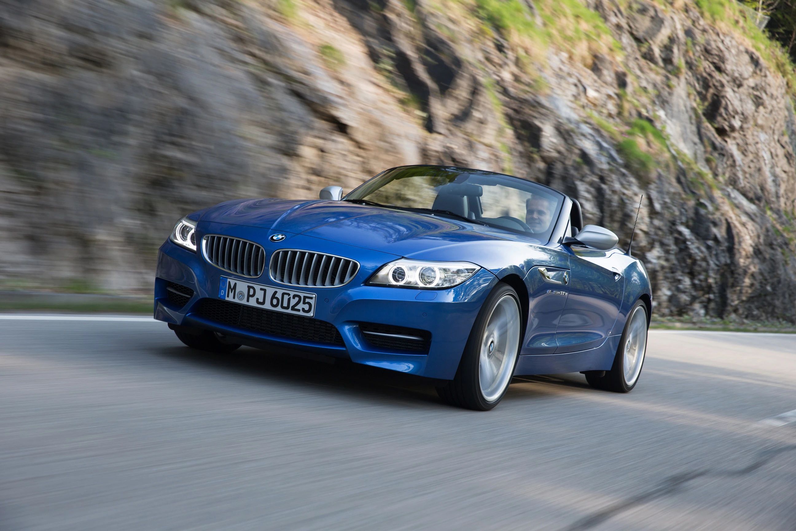 Blauer BMW Z4 Cabrio fährt dynamisch auf einer kurvigen Landstraße vor einer felsigen Kulisse.