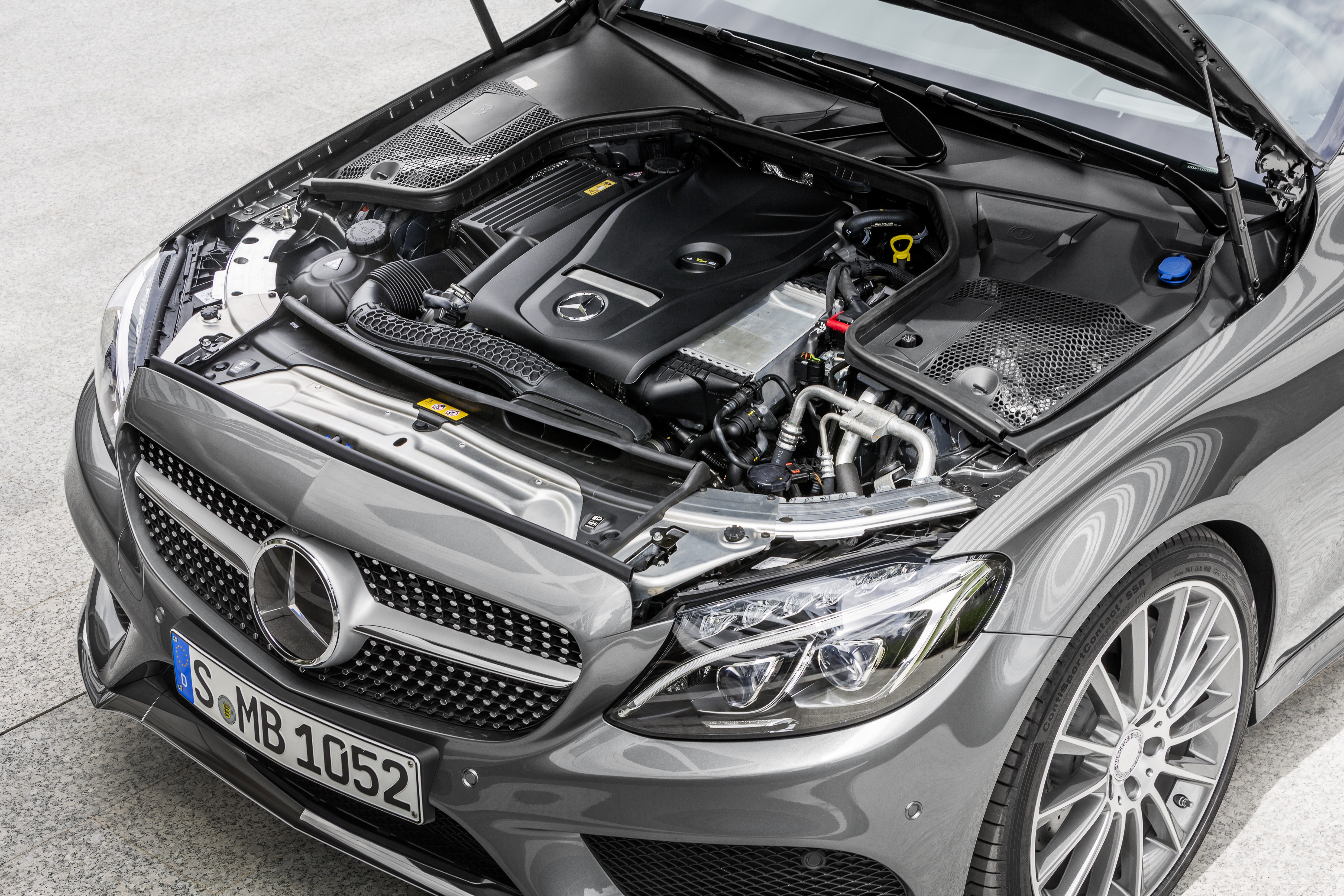 Motorraum eines grauen Mercedes-Benz, geöffnet. Sicht auf den Motorblock und verschiedene Autoteile. Vorderes Nummernschild: S-MB 1052.