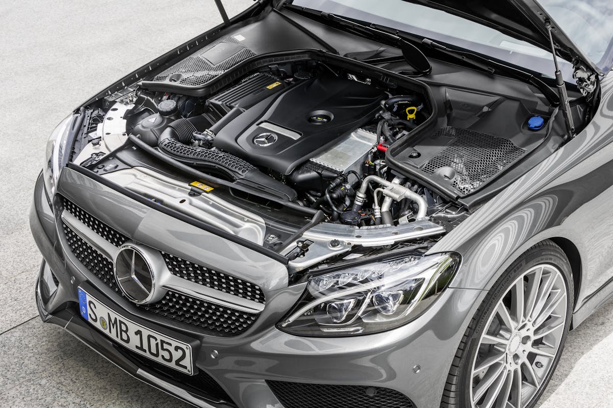 Motorraum eines grauen Mercedes-Benz, geöffnet. Sicht auf den Motorblock und verschiedene Autoteile. Vorderes Nummernschild: S-MB 1052.
