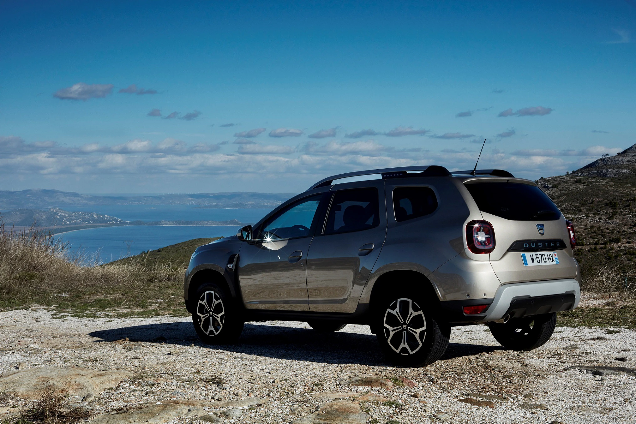 Ein silberner SUV parkt auf einer Anhöhe mit Blick auf eine Küstenlandschaft und das Meer unter einem blauen Himmel.