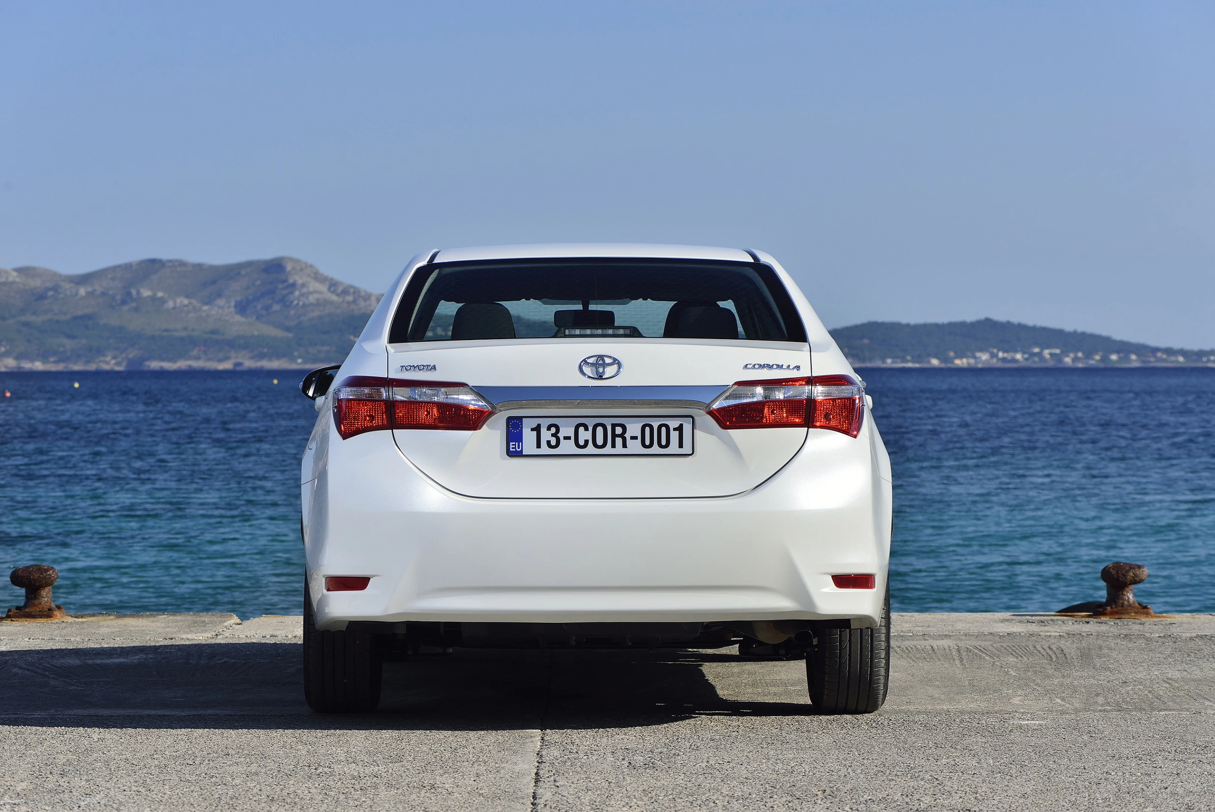Weißer Toyota Corolla steht auf einem Betonpier, mit Blick auf das Meer und eine Berglandschaft im Hintergrund.