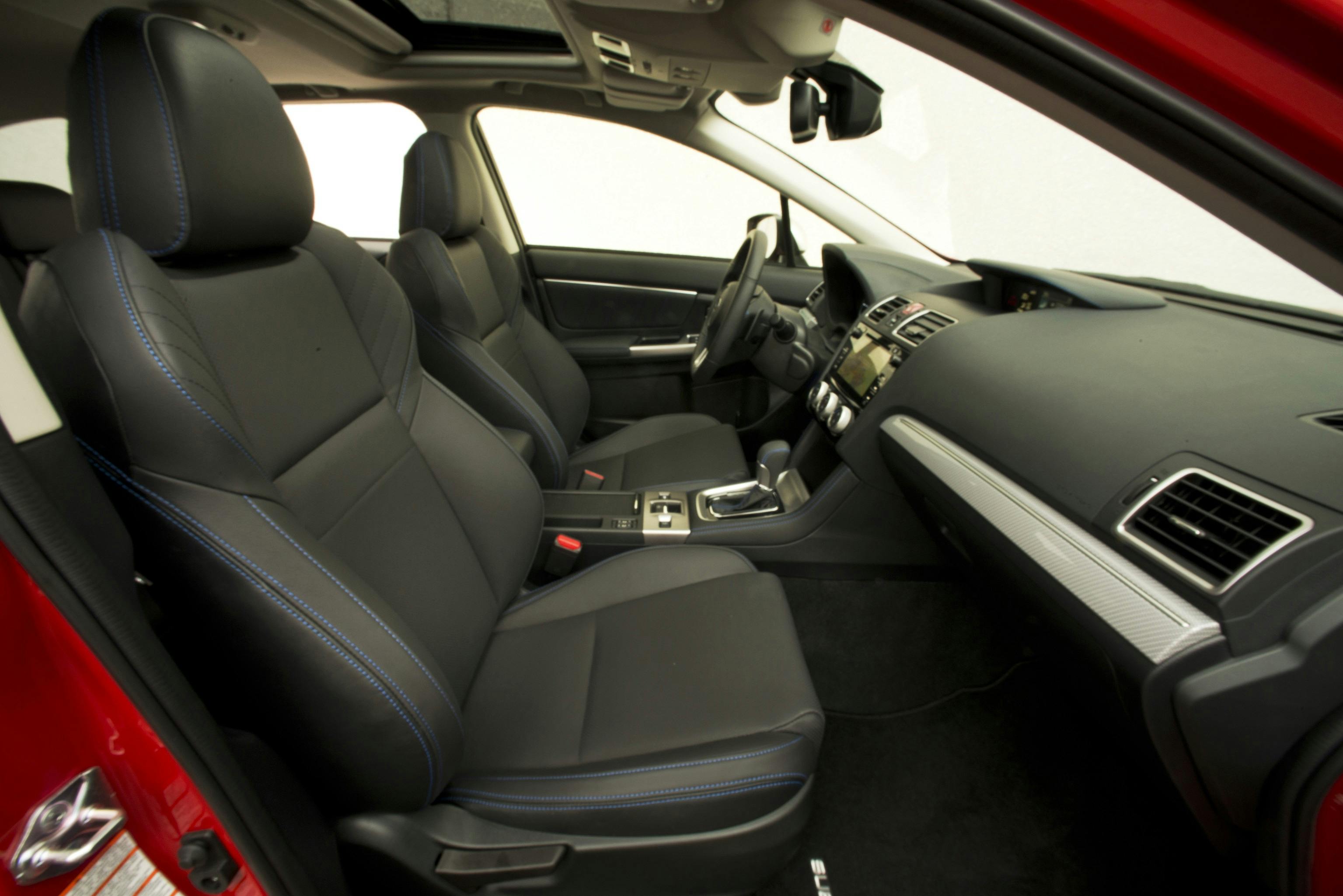 Innenraum eines Autos mit schwarzen Ledersitzen, blauen Nähten, roter Außentür und Schiebedach. Schalthebel und Armaturenbrett sind sichtbar.