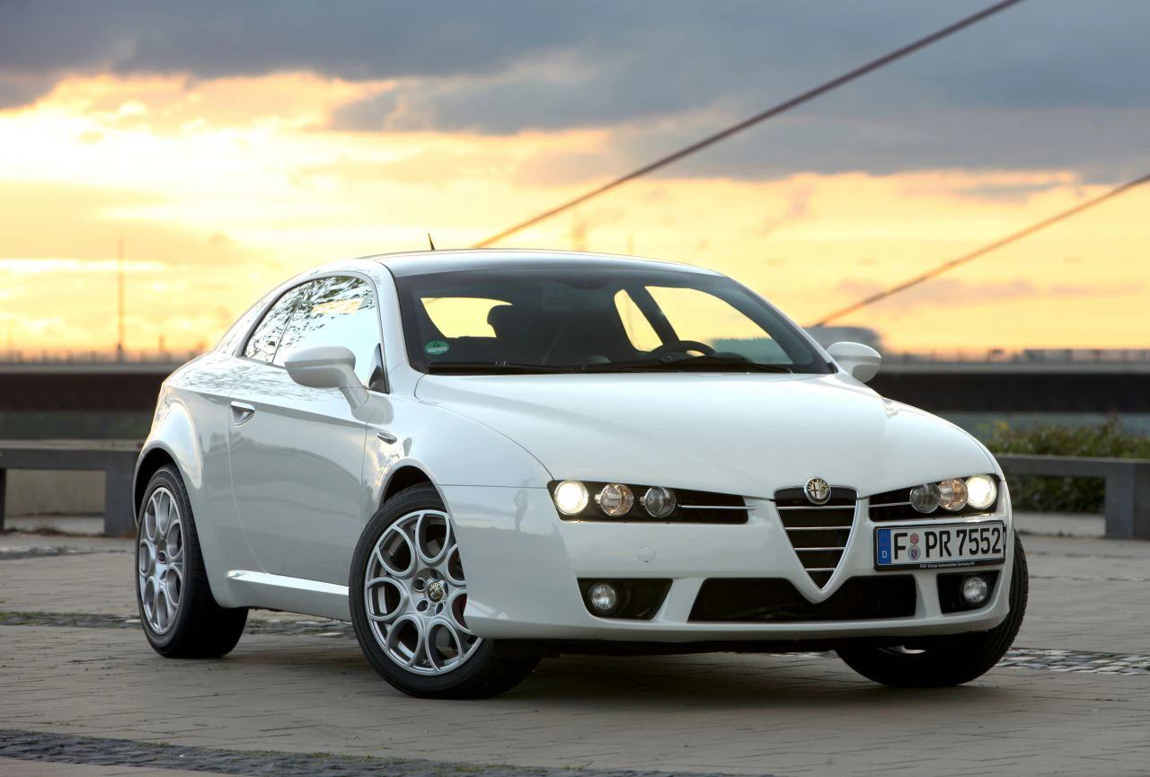 Weißer Sportwagen Alfa Romeo Brera, aufgenommen bei Sonnenuntergang auf einer Straße mit modernem Brückendesign im Hintergrund.
